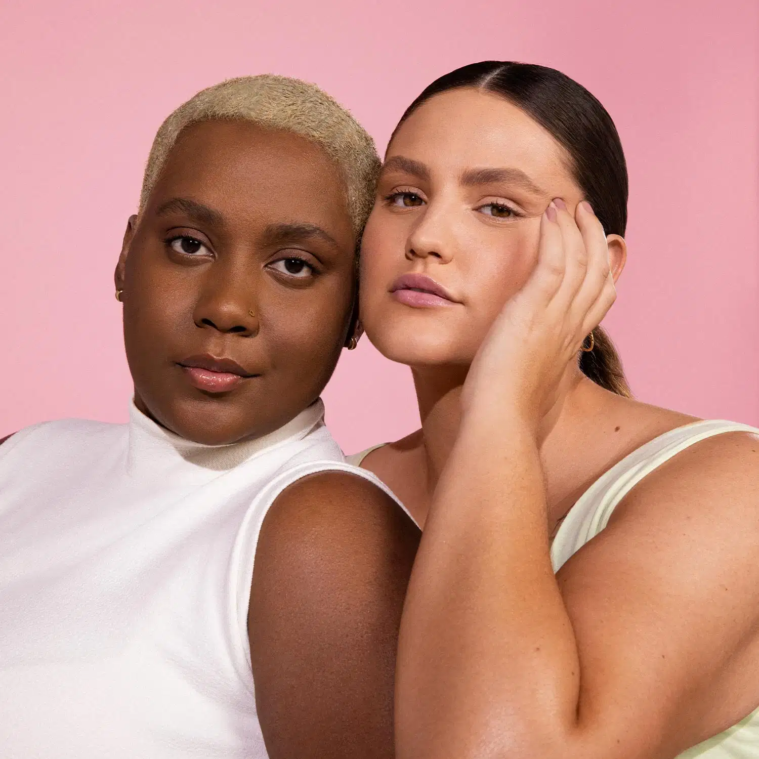 Uma mulher de pele negra e uma mulher de pele branca juntas, as duas usando roupas brancas e maquiagens leves, sobre fundo lilás.