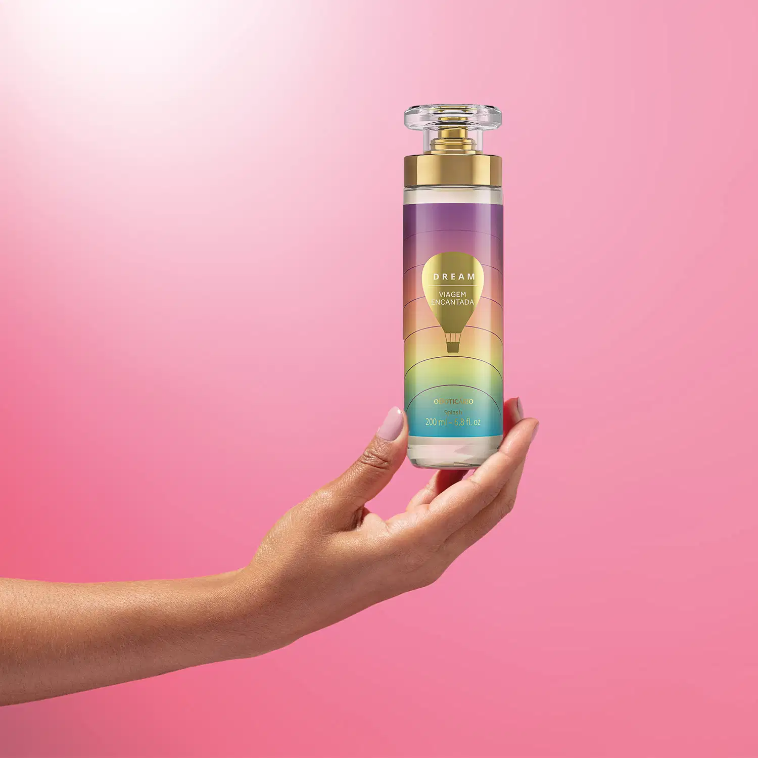 Imagem de uma mão segurando um frasco de body splash Dream Viagem Encantada. A embalagem conta com tampa transparente, válvula spray dourada e rotulo em tons de roxo, rosa e azul com balão ilustrativo. O cenário traz um fundo em tons de lilás e branco.