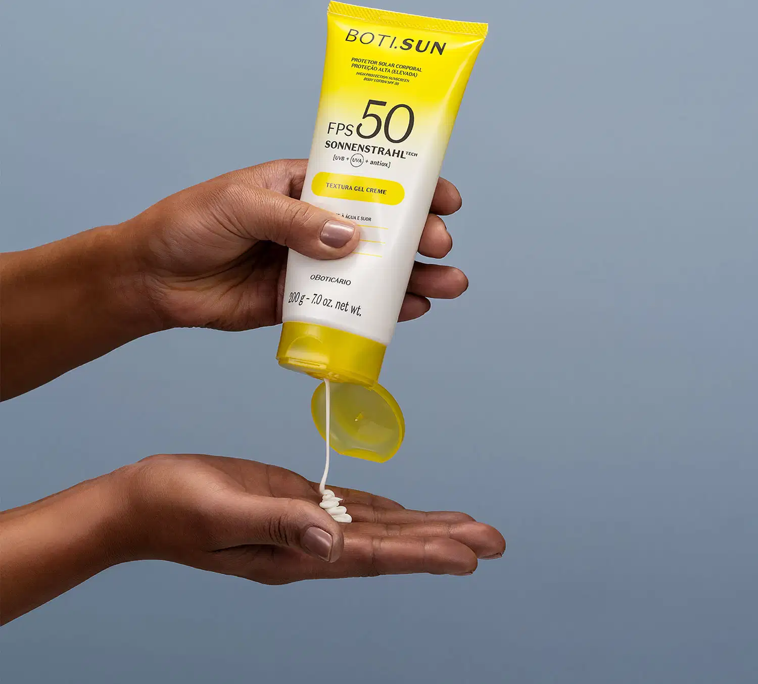 Mão segurando uma Bisnaga amarela e branca com tampa amarela e letras pretas contendo protetor solar corporal FPS 50 da marca Boti.Sun e jogando um pouco do creme branco na outra mão em fundo azul celeste.