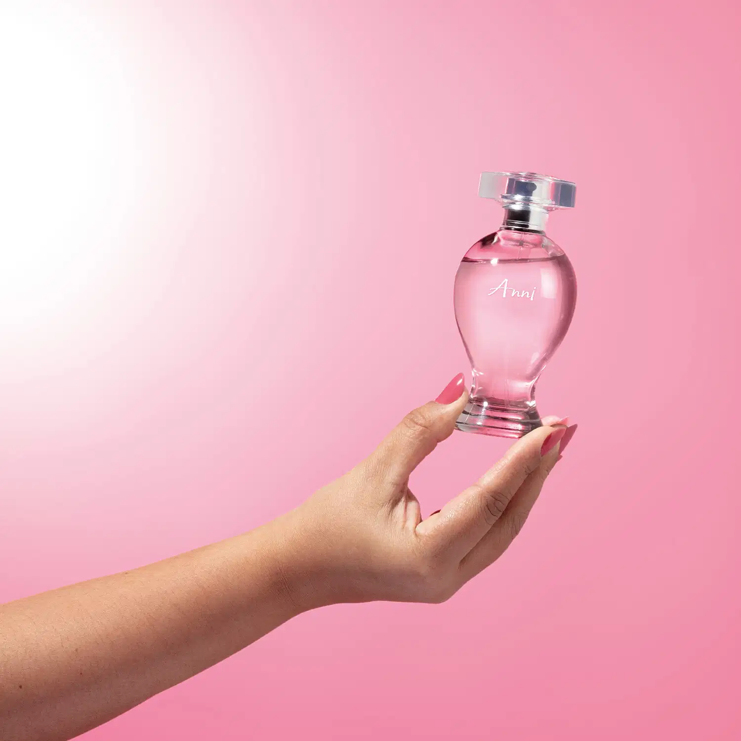Mão feminina segurando um vidro curvilíneo transparente com liquido rosa, pulverizador prateado e tampa cristal contendo desodorante colônia Boticollection Anni em fundo rosa.
