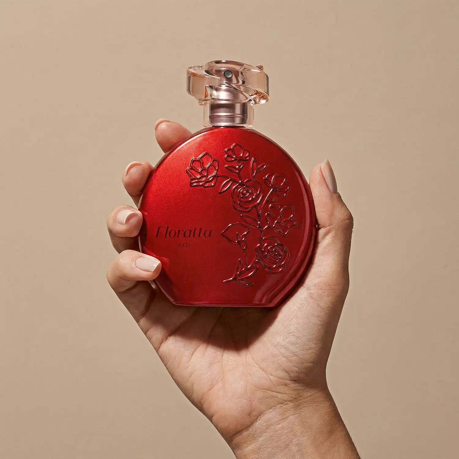 Frasco de item de perfumaria vermelho com relevo floral da linha Floratta Red sendo segurado por mãos femininas em fundo bege.