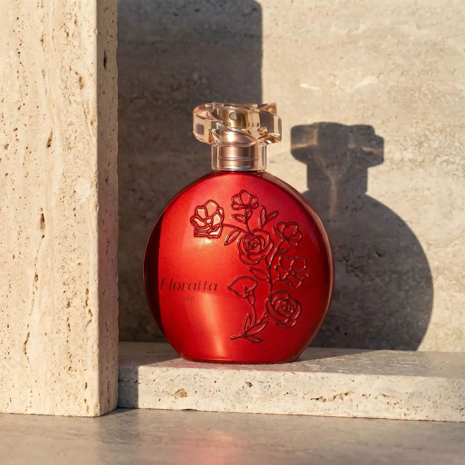 Frasco de item de perfumaria da marca Floratta Red na cor vermelha com relevo floral, apoiado sobre superfície de pedra clara, com sombras projetadas ao fundo criada por luz natural.
