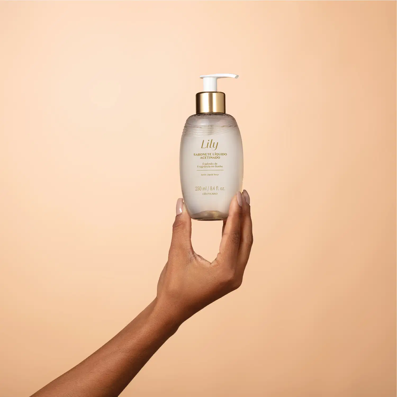 Uma mão feminina segura um frasco ovalado bege com letras marrons, detalhe dourado e bomba dosadora branca de sabonete líquido acetinado da marca Lily em um cenário com fundo em tons de bege.
