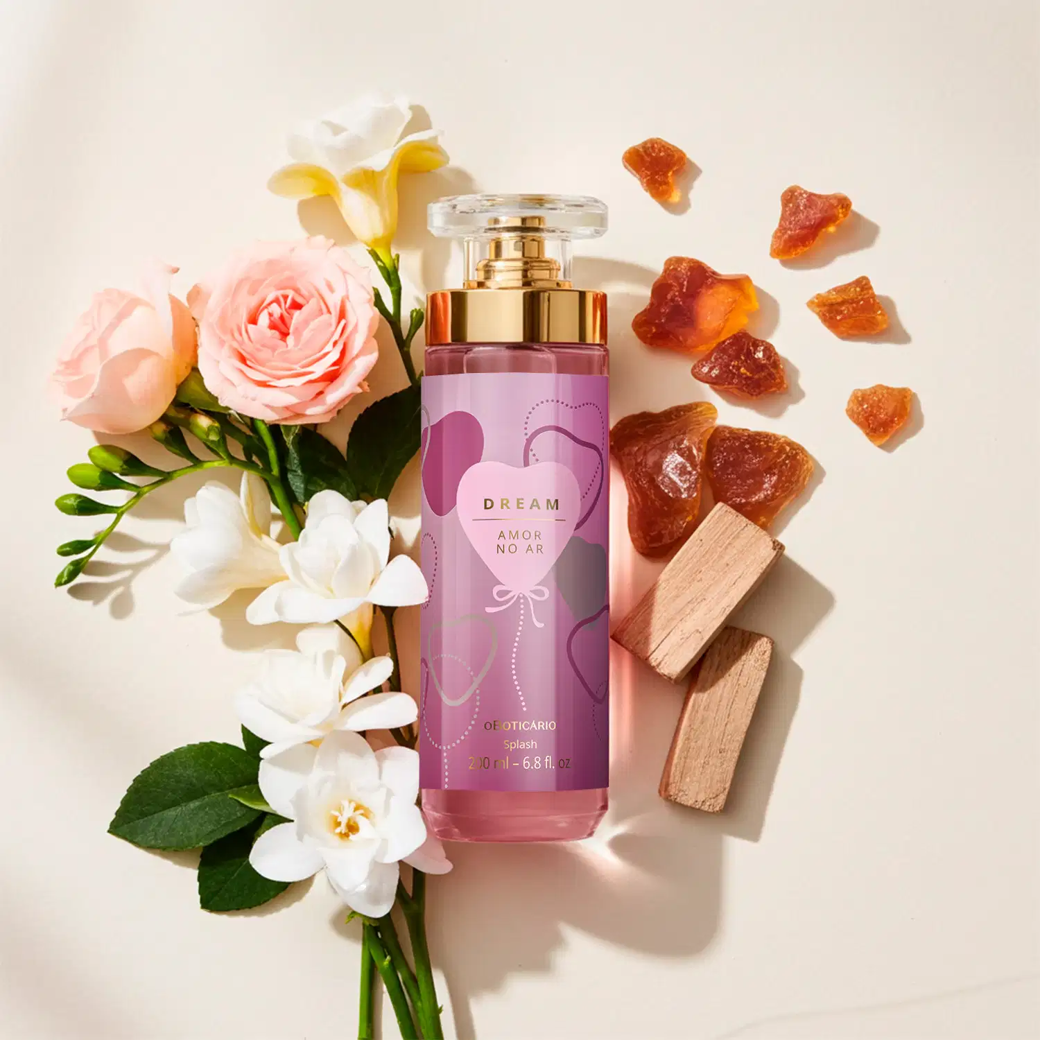 Imagem de um arranjo floral com rosas, orquídeas e folhagens verdes em fundo claro. Ao lado, frasco de body splash Dream Amor no Ar, com tampa transparente e válvula spray dourada. O rótulo é em tons de roxo e tem um coração estampado.