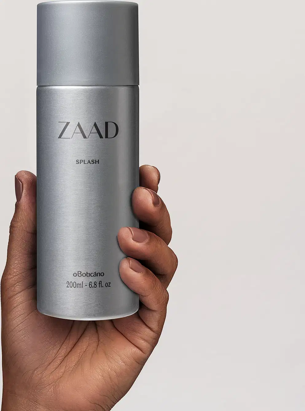 Mão segurando um frasco cilíndrico prateado com tampa e letras cinzas contendo 200ml de body splash Zaad em fundo cinza claro.