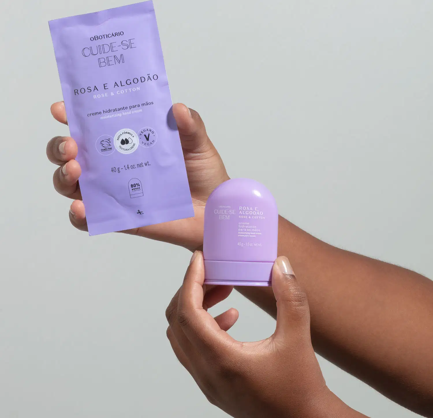 Uma mão segurando um refil de creme hidratante para mãos da marca Cuide-se Bem Rosa e Algodão em sachê de 40g na cor lilás com letras pretas e a outra segurando a embalagem original do produto na forma arredondada e na cor azul. O fundo da imagem é azul.