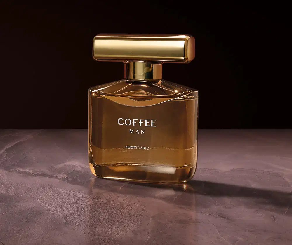 Imagem de um vidro do desodorante colônia Coffee Man em fundo escuro, com superfície de pedra e iluminação dourada. A embalagem possui uma luxuosa tampa com rotulo dourado.