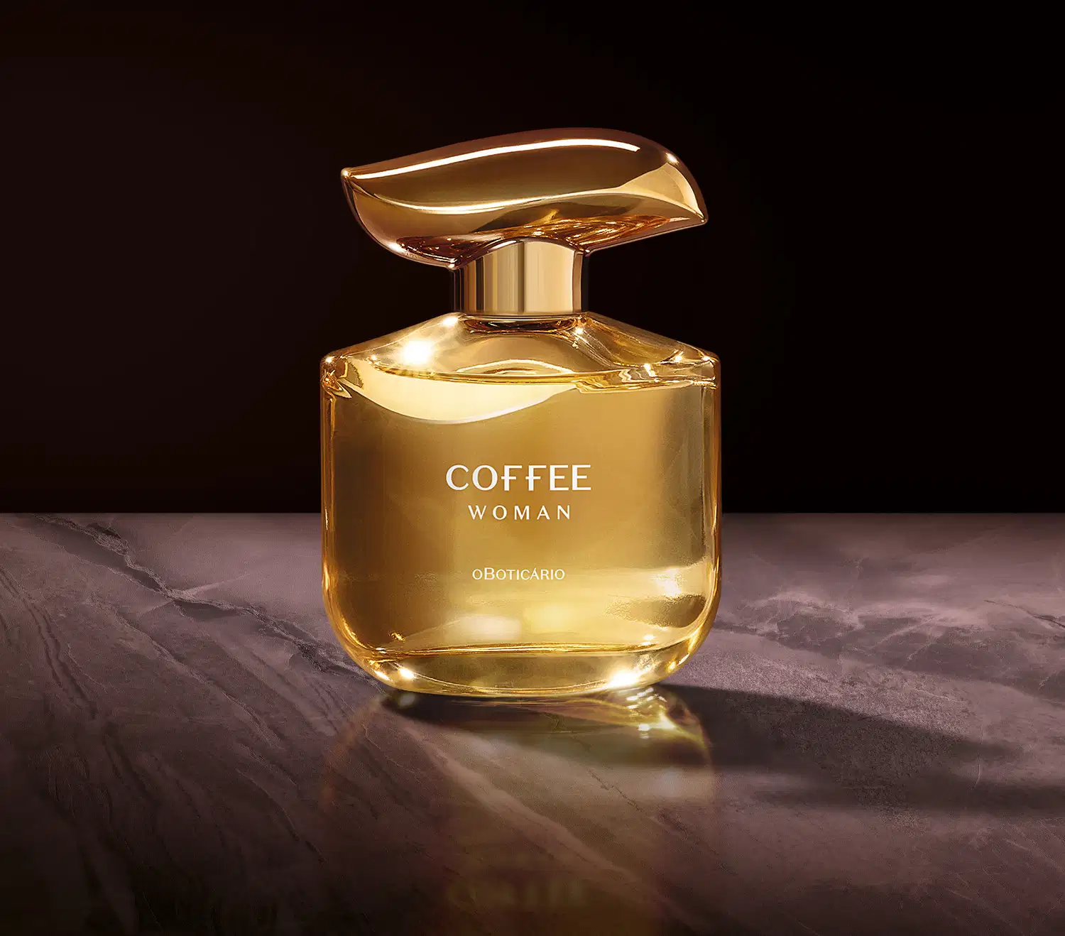 Imagem de um frasco do desodorante colônia Coffee Woman Floriental Ambarado, com cantos arredondados, em tons de dourado e marrom e letras brancas; Ele está sobre superfície escura refletiva, com iluminação suave e ambiente escuro ao redor.
