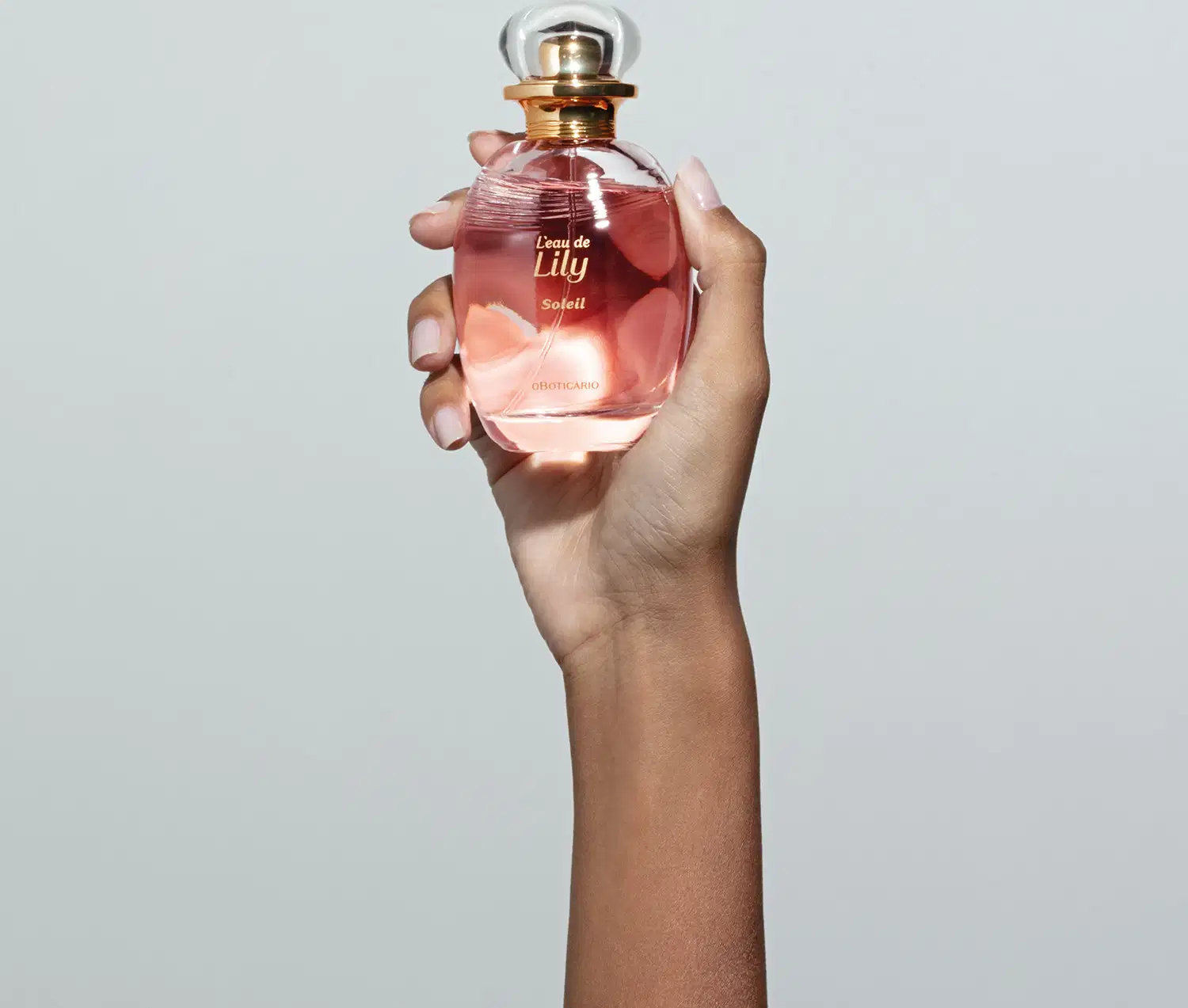 Mão feminina segurando um vidro transparente oval com líquido rosa, borrifador dourado e tampa transparente contendo desodorante colônia L'eau de Lily Soleil em fundo cinza.