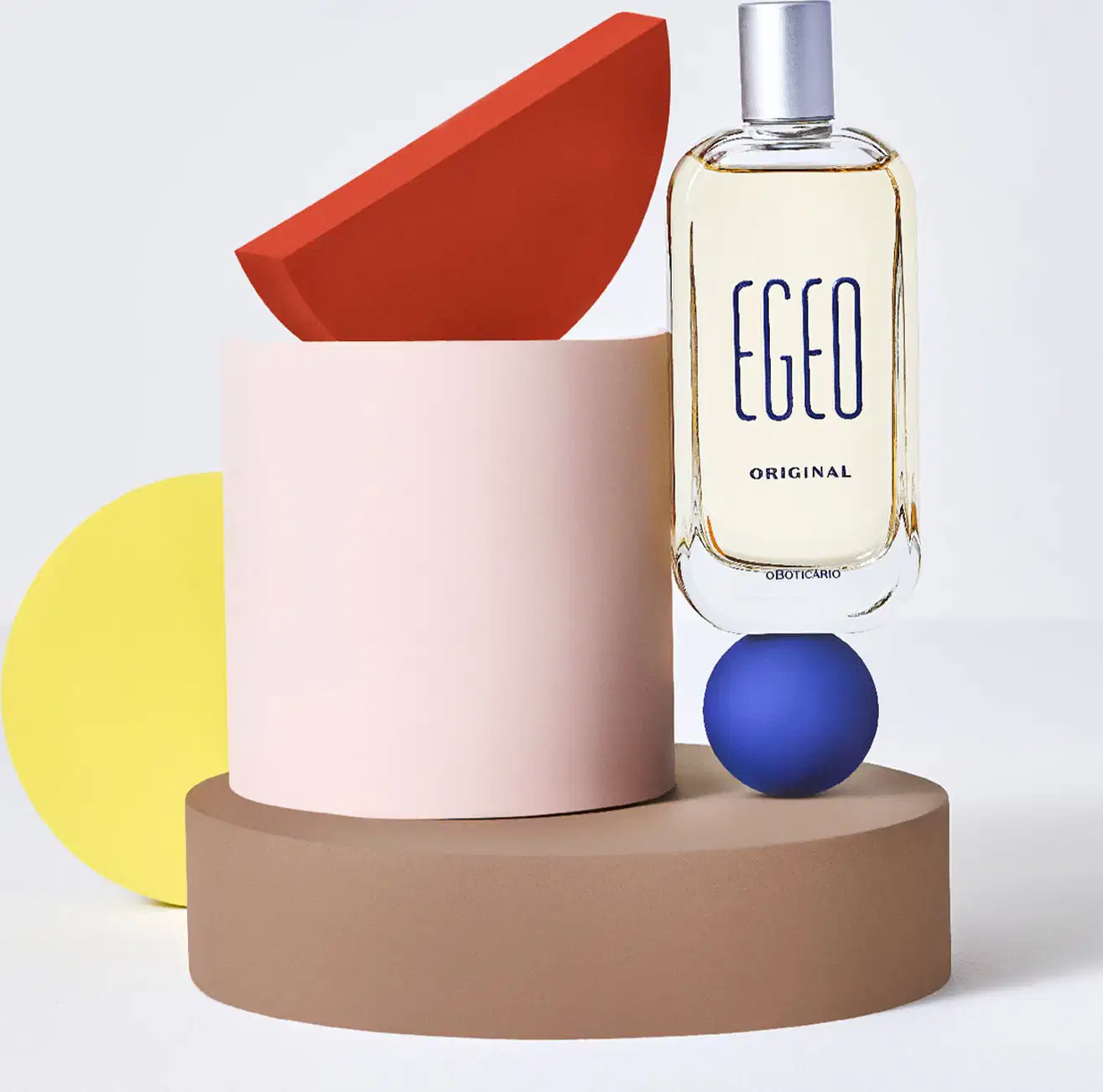 Vidro com líquido bege claro, tampa prateada e letras azul-marinho em relevo de desodorante colônia Egeo Original, posicionado em composição com cilindro rosa, formas geométricas em vermelho, amarelo e azul, sobre base circular bege em fundo cinza claro.
