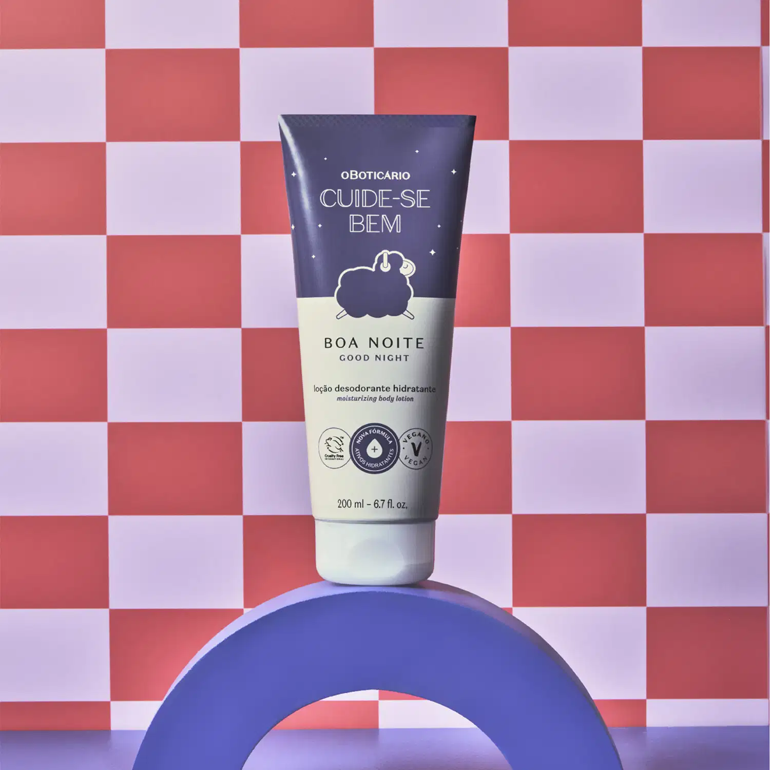 Bisnaga nas cores azul e lilás com tampa branca e ilustração de ovelhinha no rótulo contendo 200ml de loção hidratante corporal Cuide-se Bem Boa Noite em fundo xadrez vermelho e branco, apoiado sobre superfície azul.