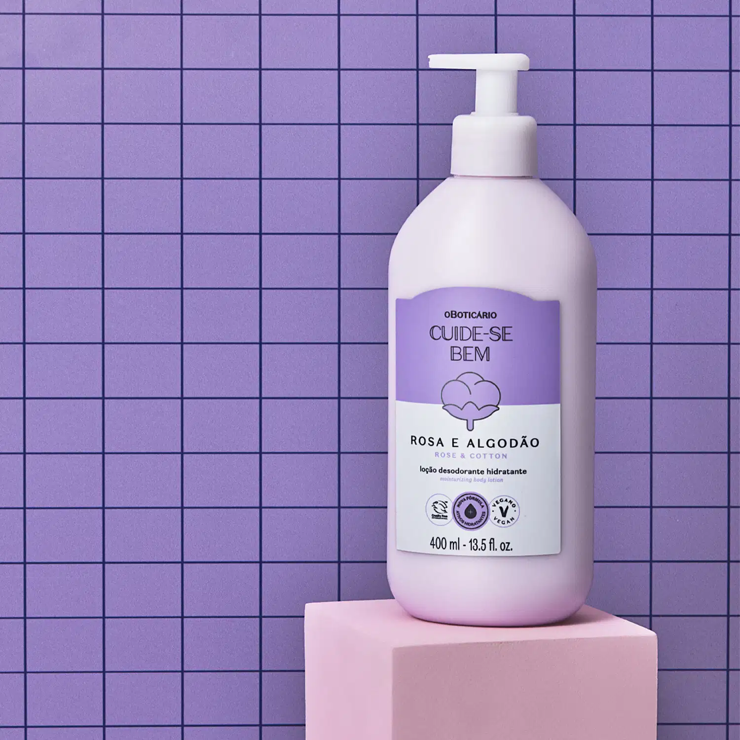 Frasco de 400ml na cor branca com rótulo lilás e branco com ilustração de uma flor de algodão contendo loção hidratante corporal Cuide-se Bem Rosa e Algodão sobre plataforma bege e fundo azul quadriculado com linhas pretas.