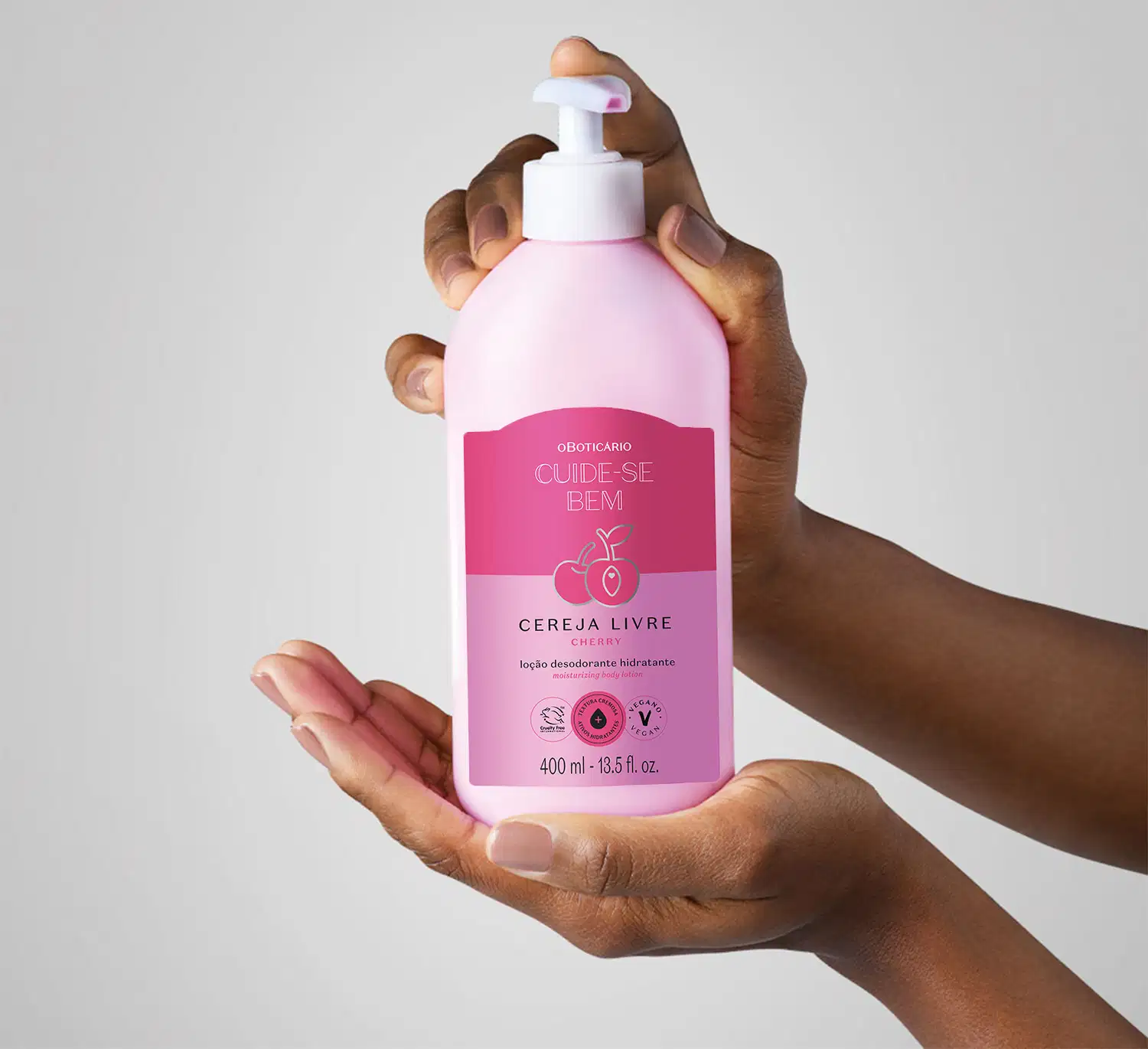 Mão feminina segurando um frasco branco com bomba dosadora e rótulo nas cores rosa e lilás com cerejas ilustradas contendo loção hidratante corporal Cuide-se Bem Cereja Livre em fundo lilás claro.