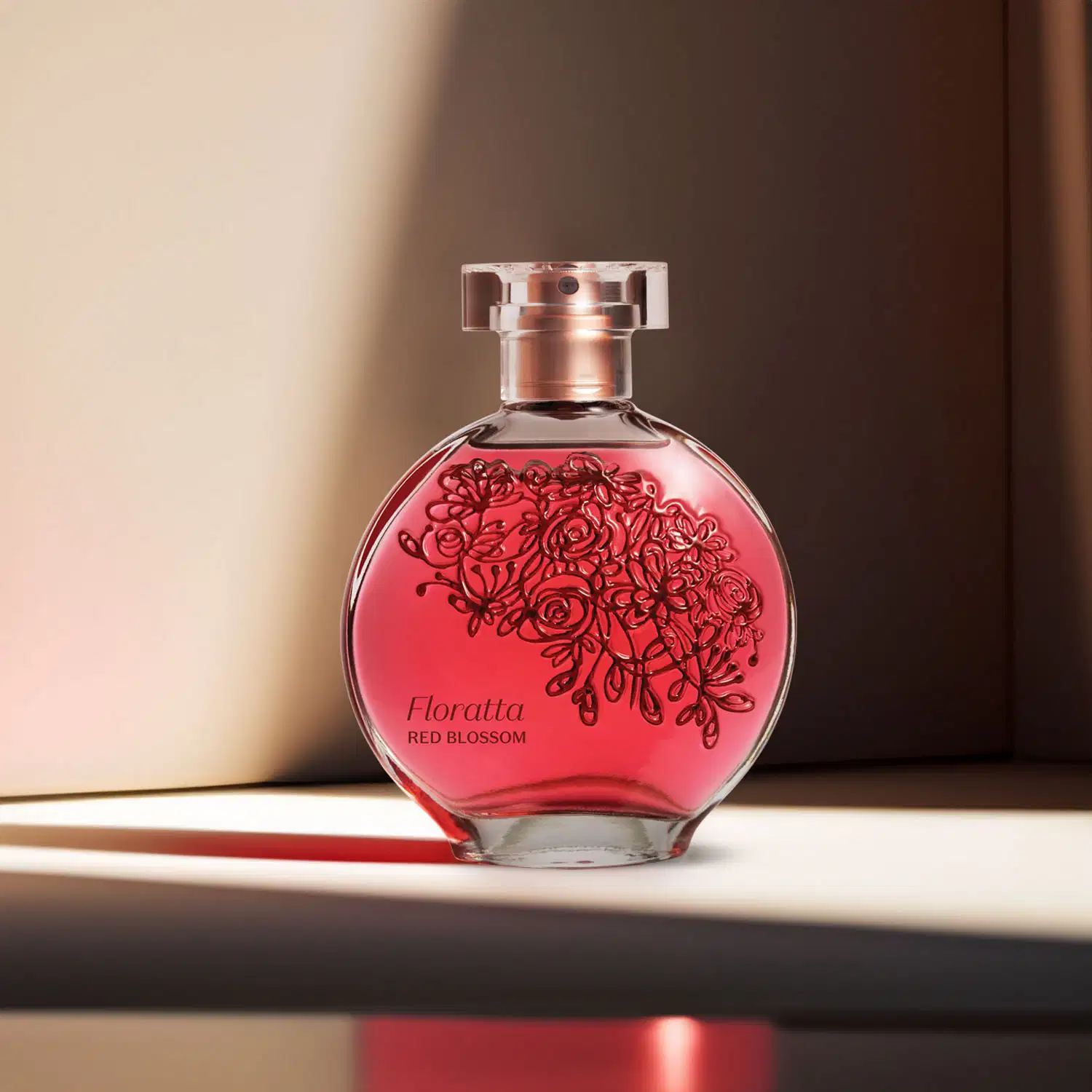 Vidro transparente com líquido vermelho, florais em relevo, borrifador dourado e tampa cristal contendo desodorante colônia Floratta Red Blossom, posicionado sobre superfície preta com sombra projetada, fundo com parede bege e marrom desfocada.