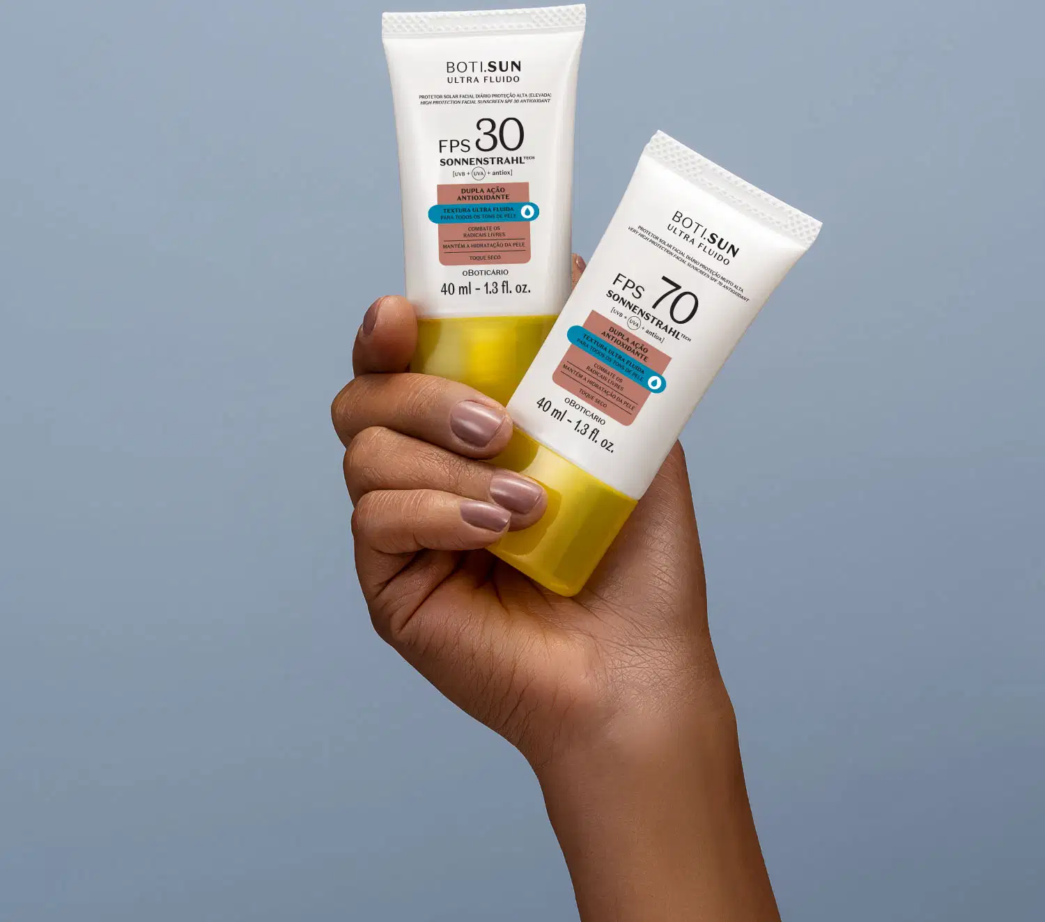Mão segurando dois frascos brancos com tampas amarelas, letras pretas e detalhes nas cores azul e laranja contendo protetor solar facial antioxidante FPS 30 Ultra Fluído da marca Boti.Sun em fundo azul celeste.