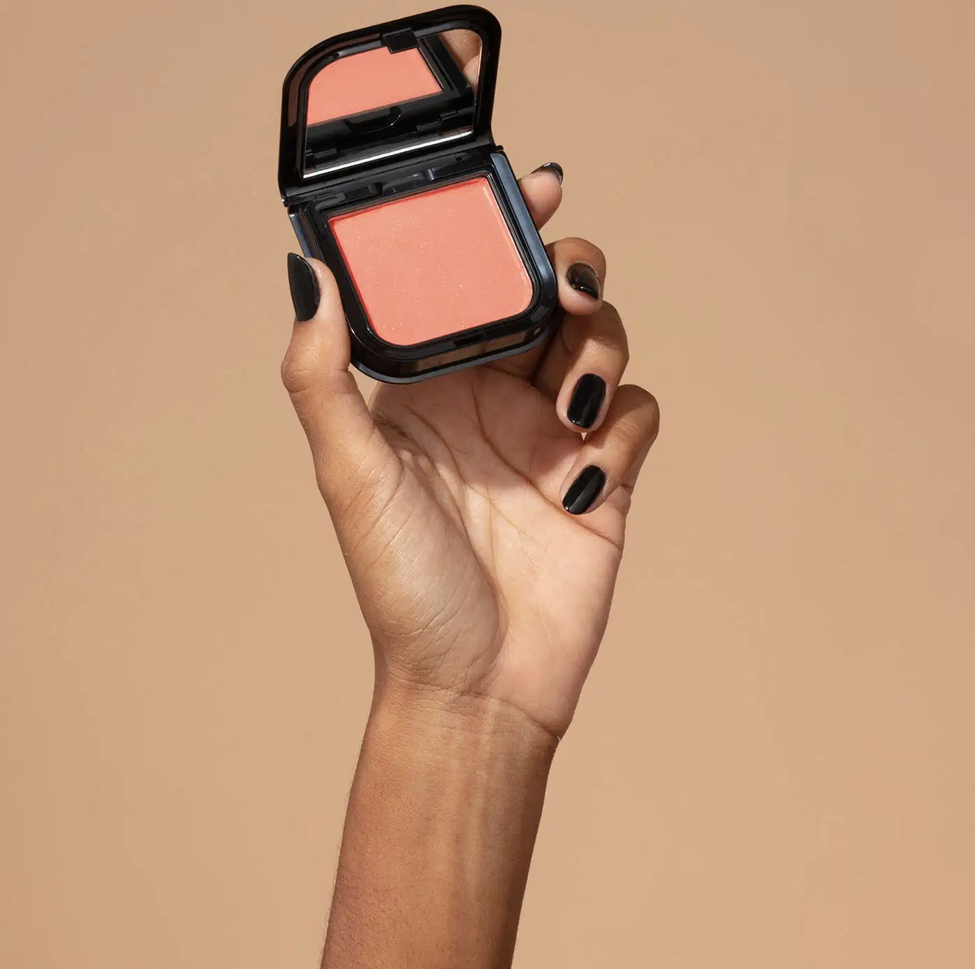 Uma mão feminina com unhas pintadas de preto está segurando um blush em pó mate Luminoso Coral Make B. na cor coral em estojo quadrado preto de cantos arredondados e espelho na tampa. O fundo da imagem é em tons de marrom.