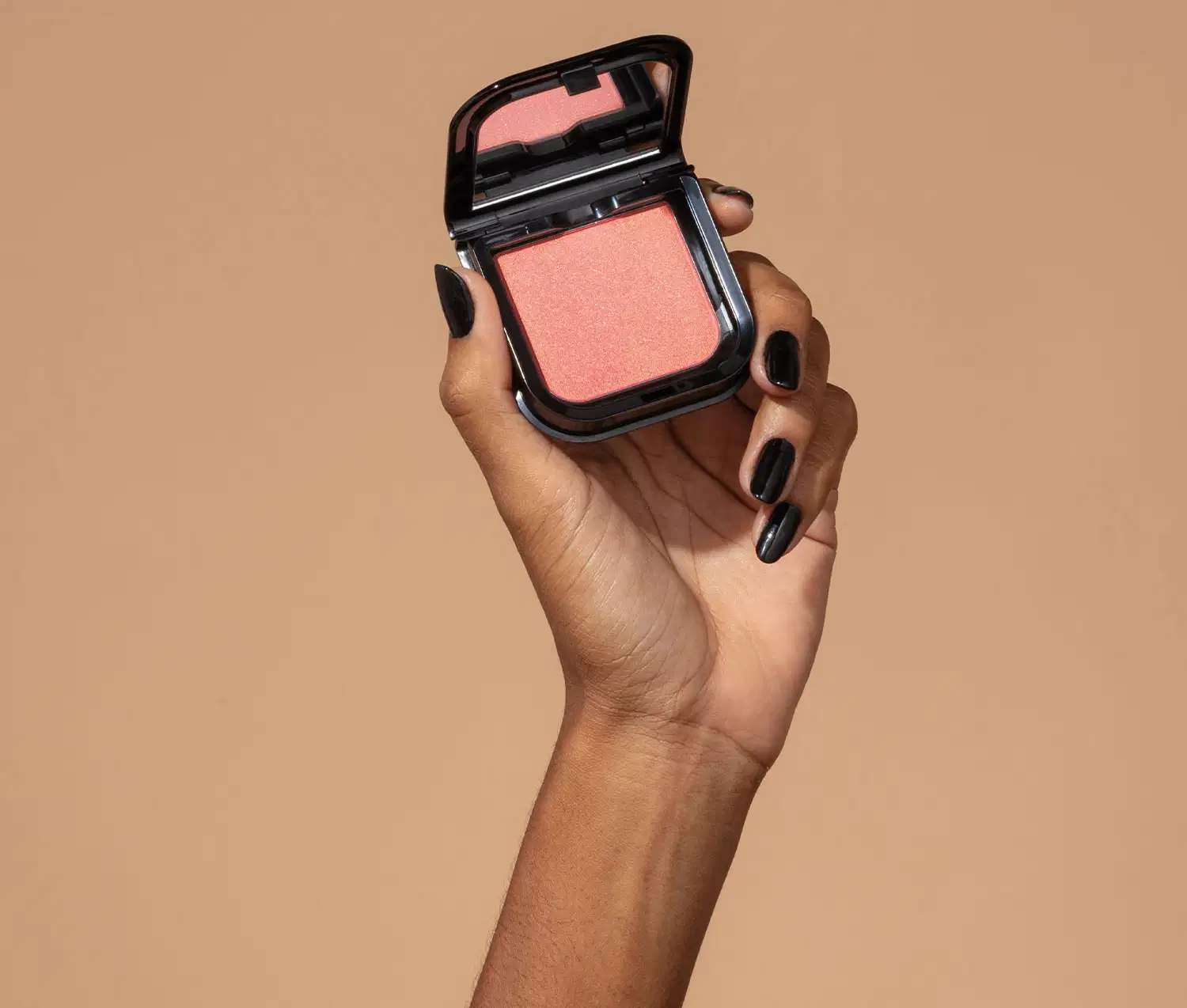 Uma mão feminina com unhas pintadas de preto está segurando um blush em pó mate Luminoso Rose Make B. na cor rosa em estojo quadrado preto de cantos arredondados e espelho na tampa. O fundo da imagem é em tons de marrom.