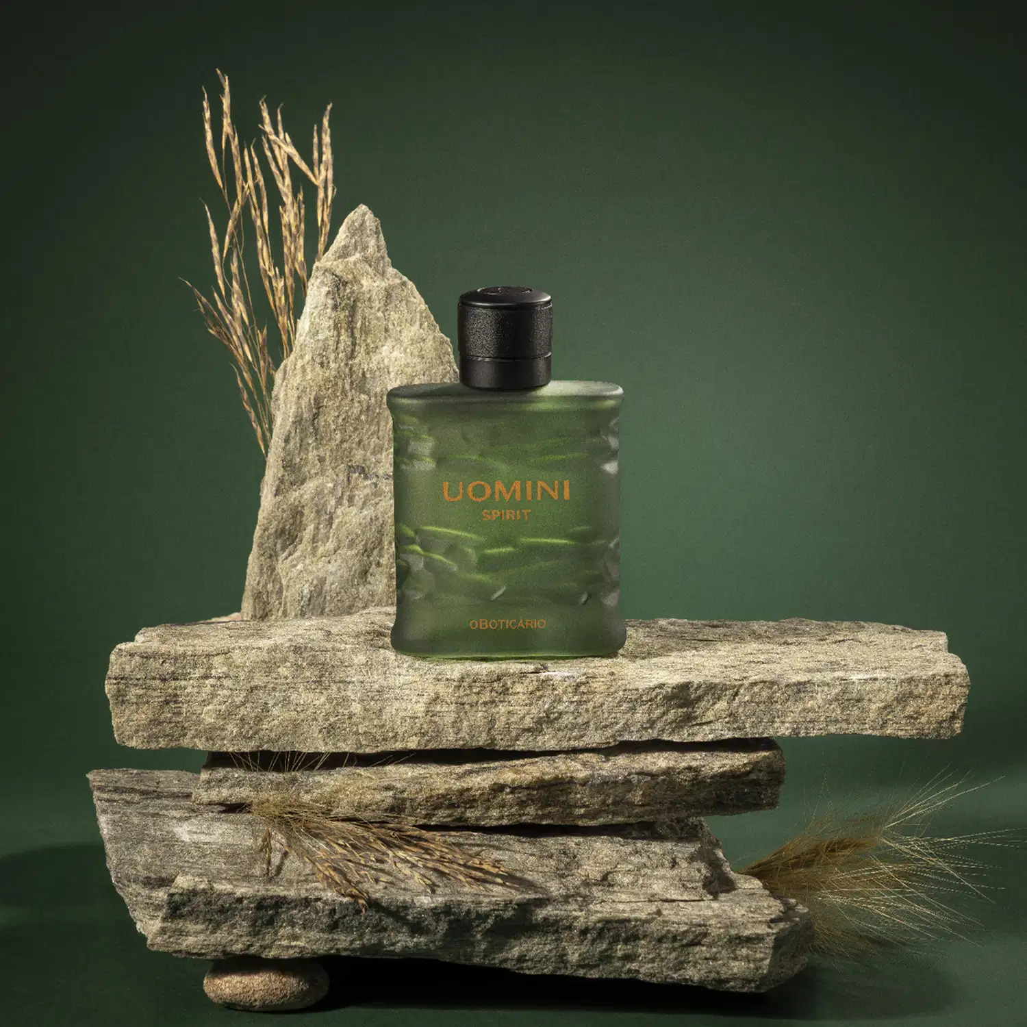 Frasco retangular em tom de verde oliva com tampa texturizada preta e letras laranjas contendo 100ml de desodorante colônia Uomini Spirit sobre superfície rochosa com galhos de trigo, em ambiente com tons de verde escuro.