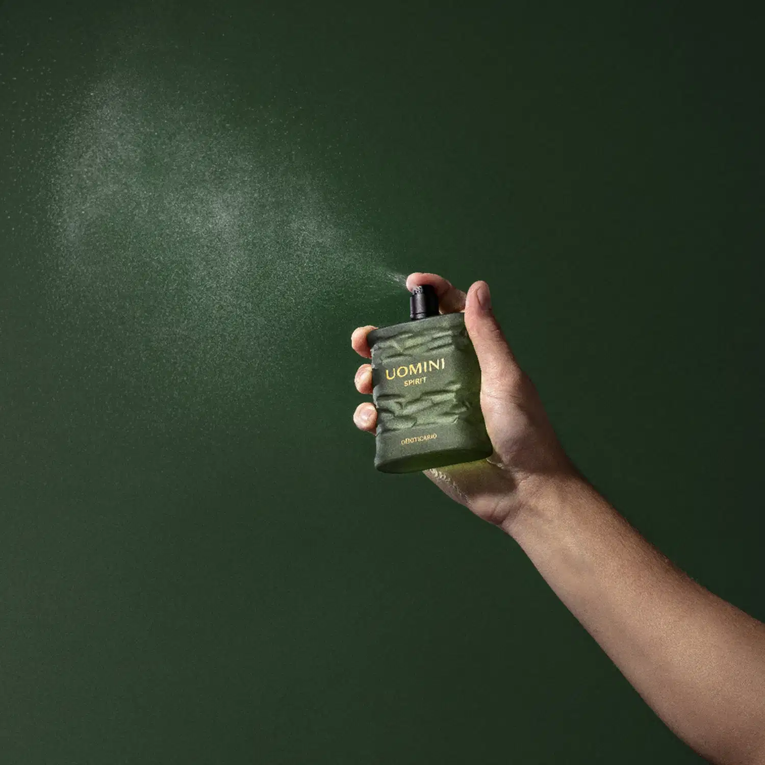 Mão segurando um frasco retangular em tom de verde oliva com tampa texturizada preta e letras laranjas contendo 100ml de desodorante colônia Uomini Spirit, espirrando o produto no ar em fundo verde bem escuro.