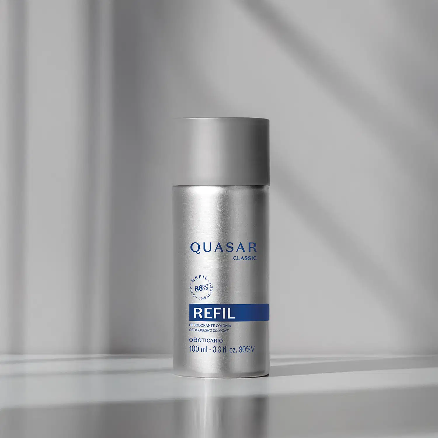 Refil de desodorante colônia Quasar Classic em frasco prateado com detalhes e letras azuis em fundo em tons de cinza com luzes naturais.