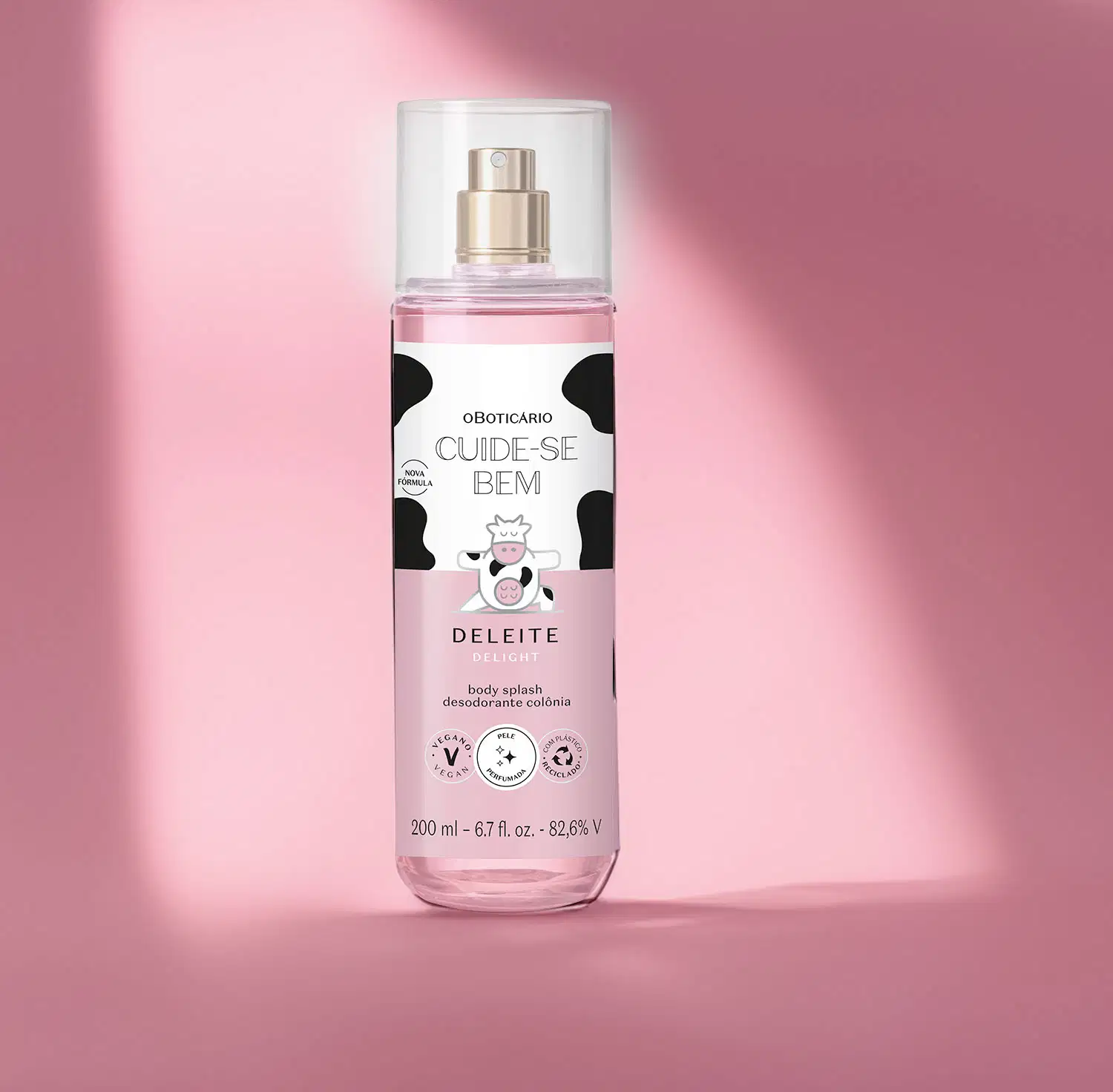 Frasco transparente com líquido rosa, pulverizador prateado, tampa transparente e rótulo branco e rosa com manchas pretas e ilustração de vaca contendo 200ml de body splash Cuide-se Bem Deleite em fundo rosa com sombras.