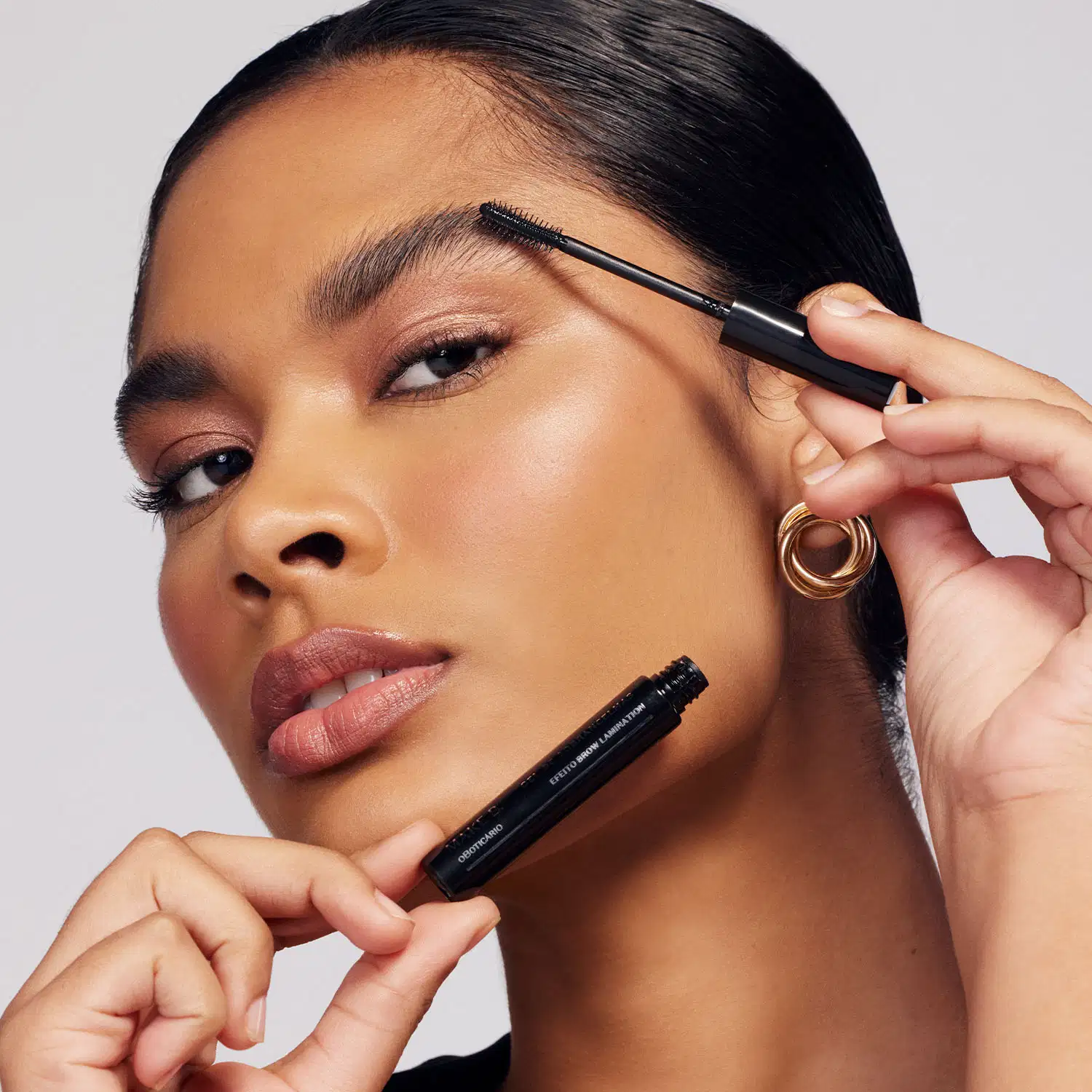 Uma mulher bem maquiada está com os cabelos presos e, em uma mão, segura um tubo preto de máscara de sobrancelhas em gel Make.B Brow Lamination e, na outra mão, segura a tampa aplicadora e aplica o produto nas sobrancelhas.