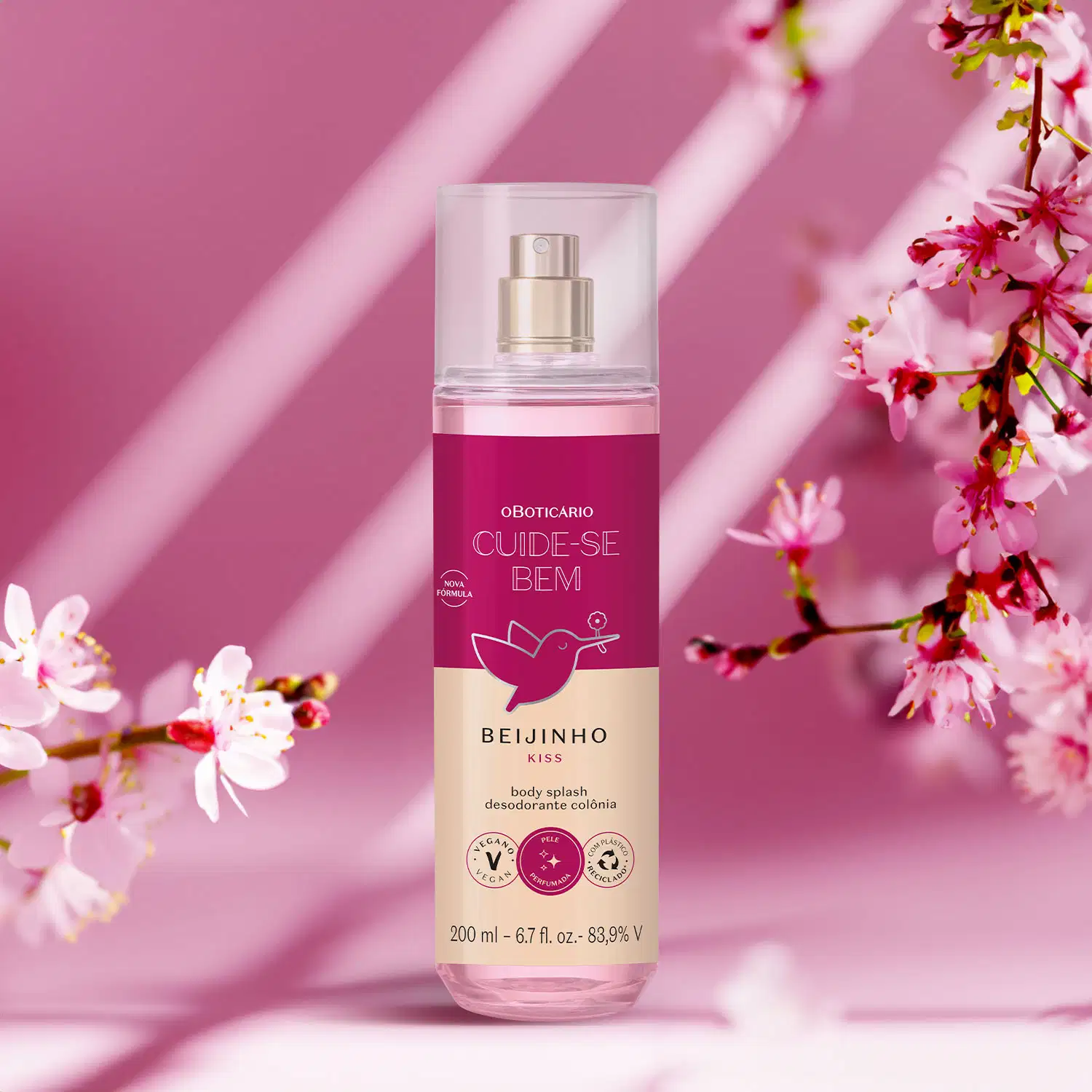 Frasco com líquido rosa, pulverizador prateado, tampa transparente e rótulo vinho e rosa com beija-flor ilustrado de body splash da marca Cuide-se Bem Beijinho com flores de cerejeira rosa ao fundo, em ambiente iluminado por raios de luz.