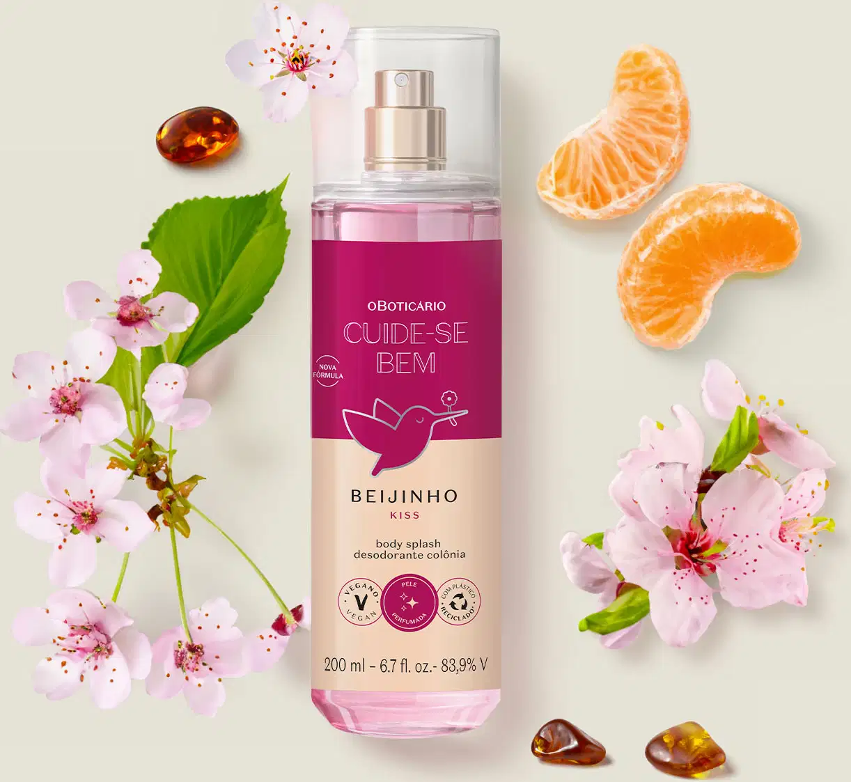 Frasco com líquido rosa, pulverizador prateado, tampa transparente e rótulo vinho e rosa com beija-flor ilustrado de body splash da marca Cuide-se Bem Beijinho. em ambiente com flores de cerejeira, fatias de laranja e pedras âmbar, sobre superfície clara.