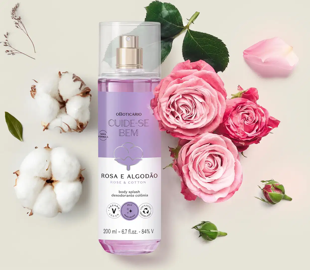 Frasco com líquido azul, pulverizador prateado, tampa transparente e rótulo azul e branco com flor de algodão ilustrada contendo body splash Cuide-se Bem Rosa e Algodão em meio a rosas cor de rosa, algodão e folhas verdes sobre superfície clara.