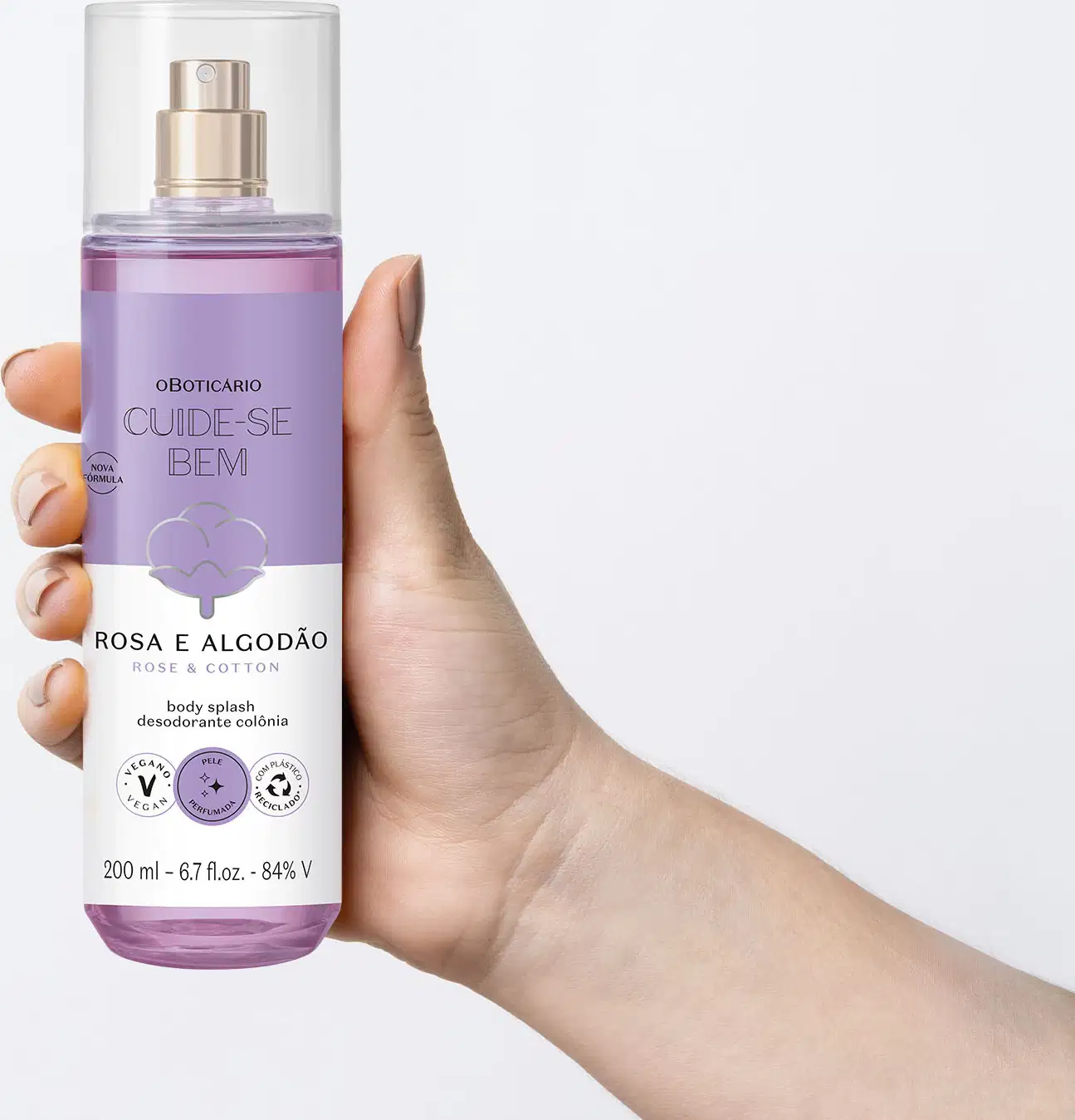 Mão feminina segurando um frasco cilíndrico com líquido azul, pulverizador prateado, tampa transparente e rótulo azul e branco com flor de algodão ilustrada contendo body splash Cuide-se Bem Rosa e Algodão em fundo azul claro.