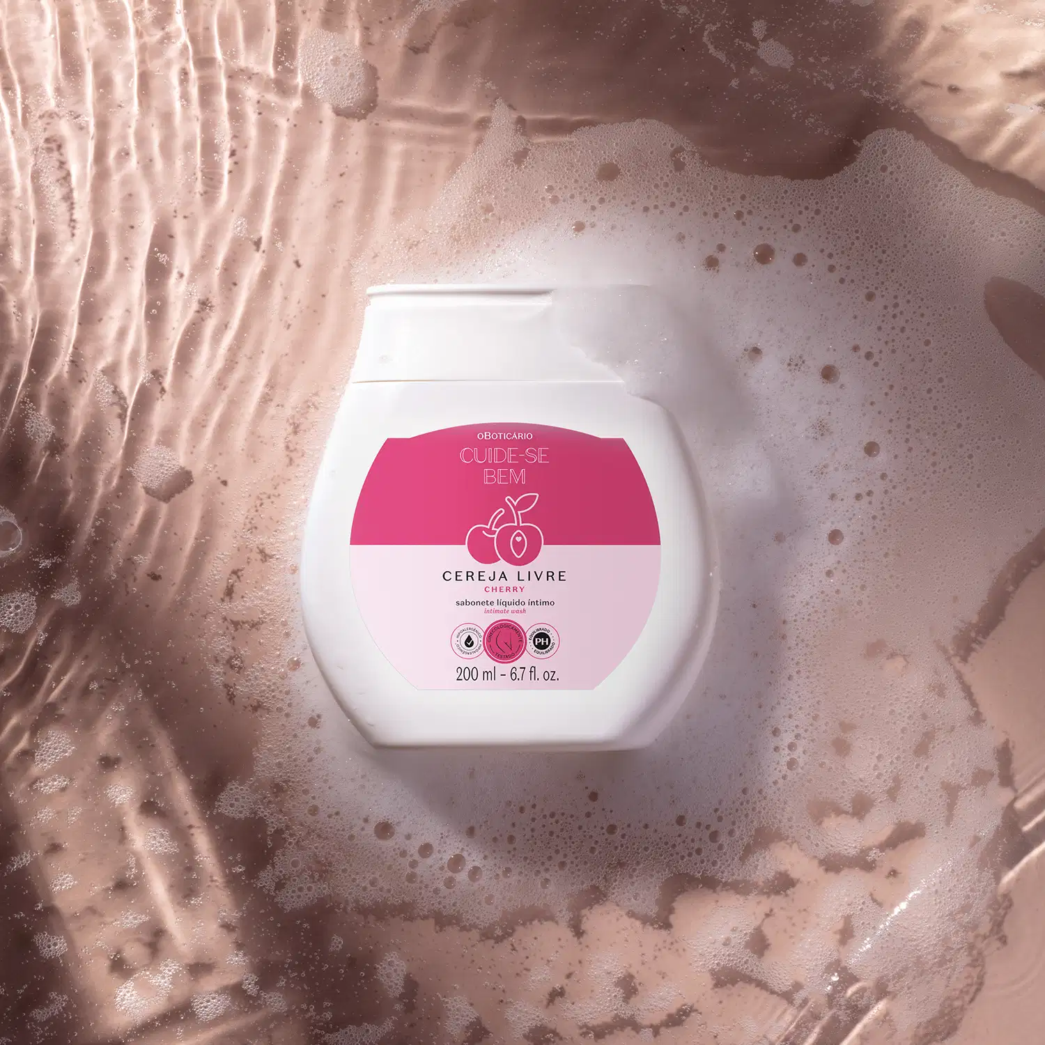 Recipiente branco com rótulo rosa da marca Cuide-se bem Cereja Livre, flutuando em espuma de banho com bolhas, em ambiente com iluminação suave e tons terrosos.