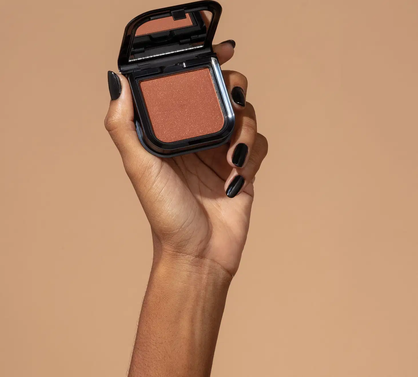 Uma mão feminina com unhas pintadas de preto está segurando um blush em pó mate Luminoso Brown Make B. na cor marrom em estojo quadrado preto de cantos arredondados e espelho na tampa. O fundo da imagem é em tons de marrom.