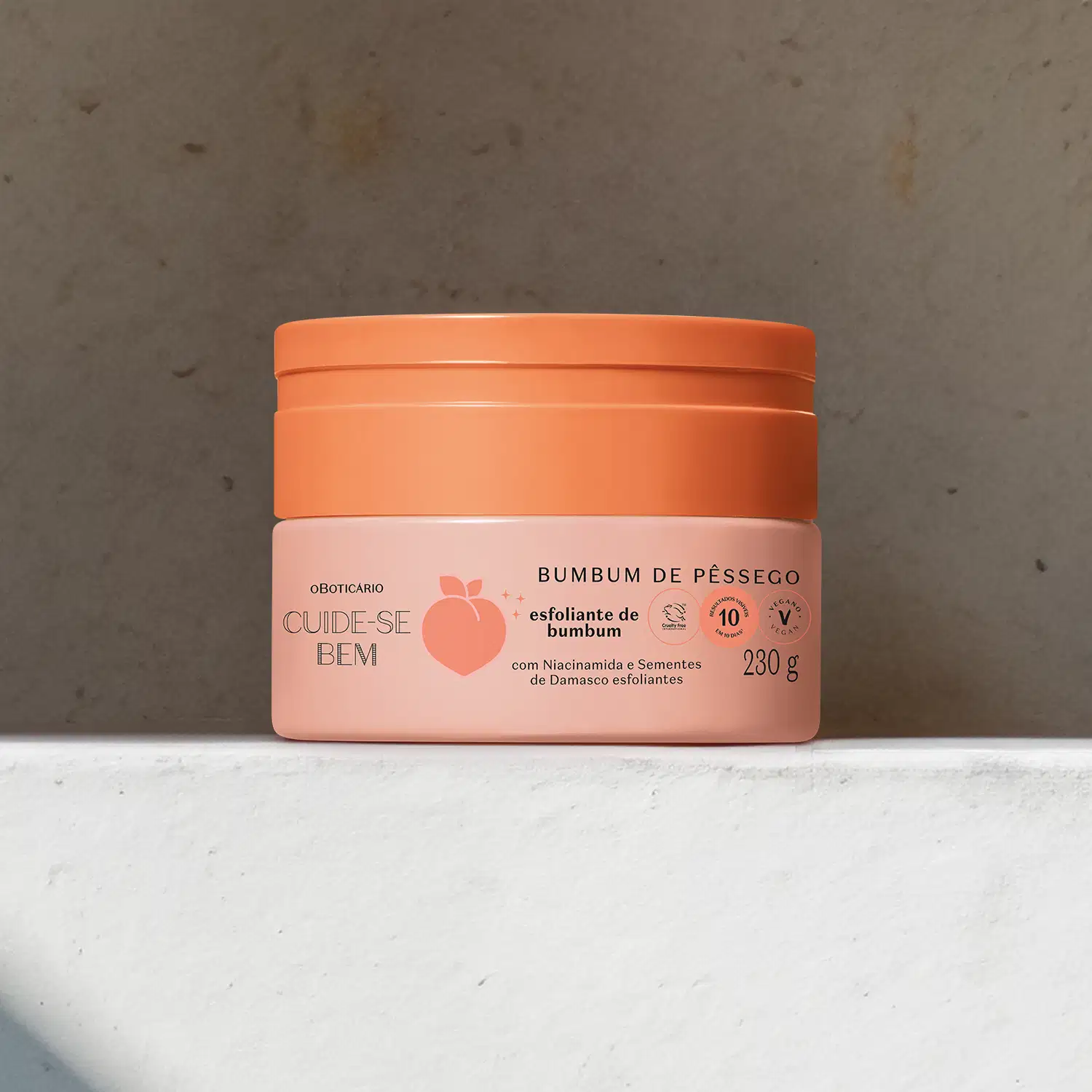 Pote em tons de rosa com letras pretas e pêssego ilustrado contendo creme esfoliante de bumbum Cuide-se Bem Bumbum de Pêssego em superfície clara e fundo marrom.