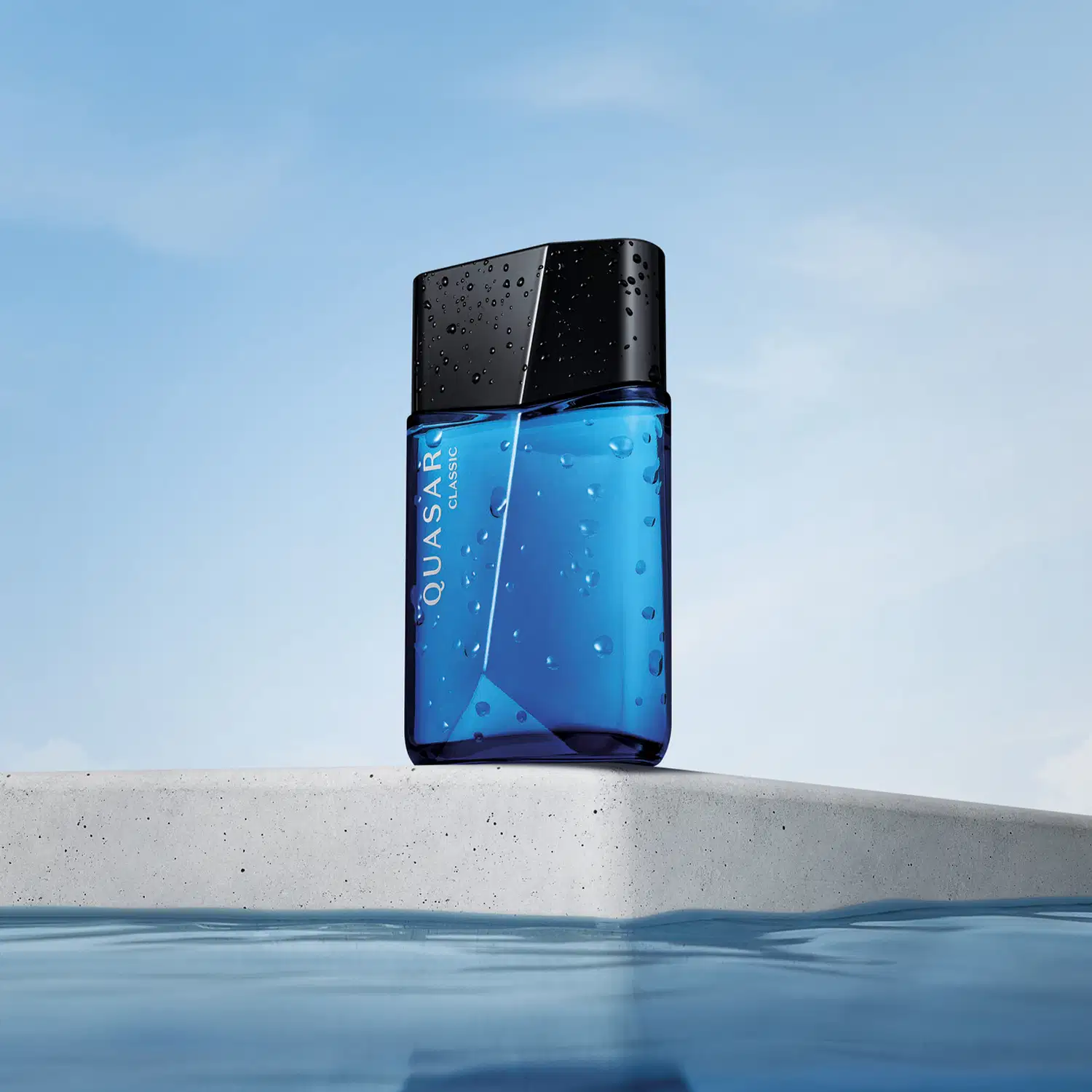 Vidro azul com tampa preta, ambos em formato geométrico e letras brancas no rótulo contendo 100ml de desodorante colônia Quasar Classic flutuando sobre superfície de concreto com água ao redor, sob céu azul claro.