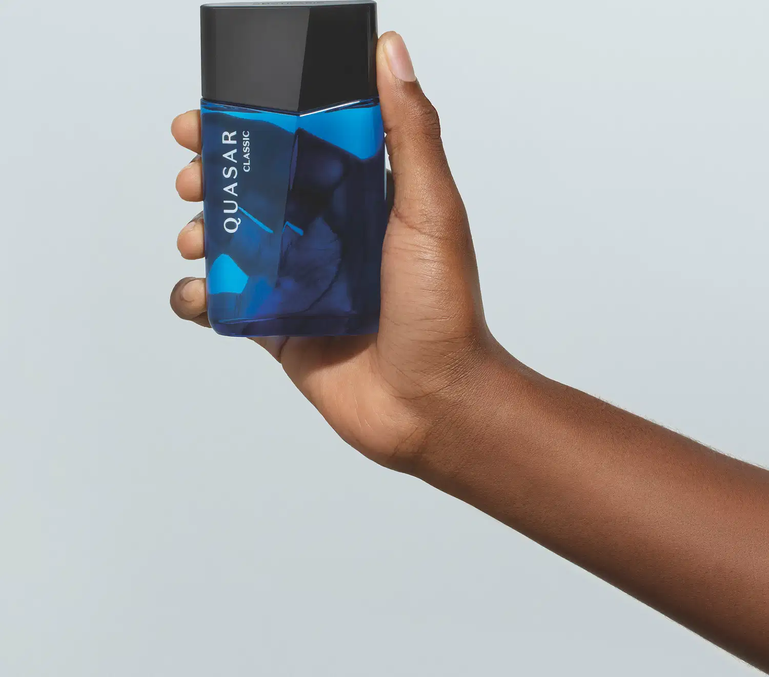 Mão masculina de pele negra segurando um vidro azul com tampa preta, ambos em formato geométrico e letras brancas no rótulo contendo 100ml de desodorante colônia Quasar Classic em fundo azul.