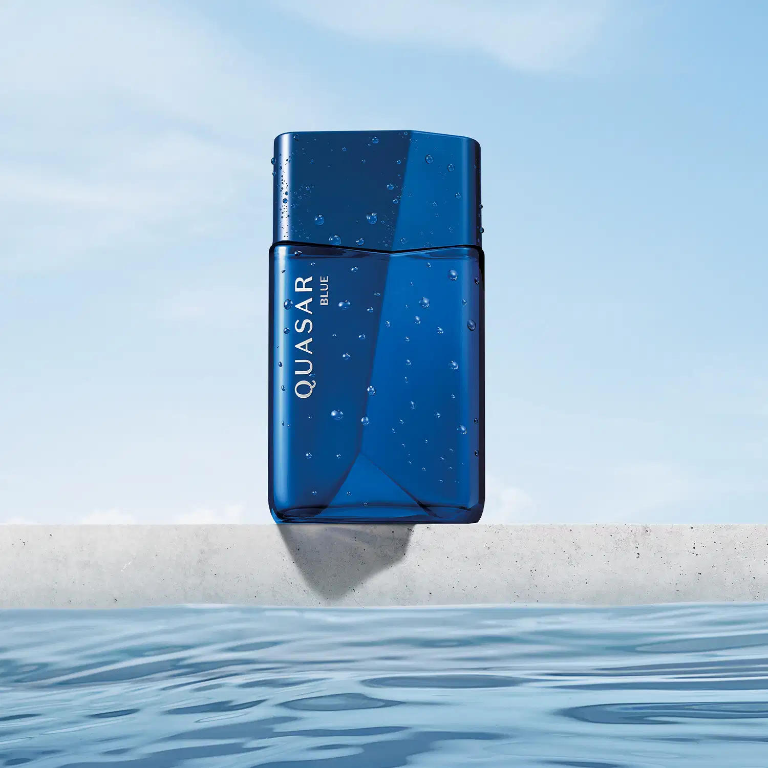 Frasco de vidro em formato geométrico com letras amarelas contendo desodorante colônia Quasar Blue com gotas d'água, flutuando sobre superfície de água em ambiente externo com céu claro.