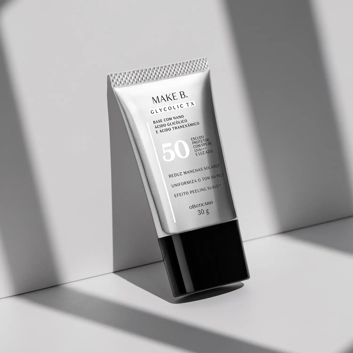 Tubo prateado com tampa e letras pretas da base líquida Make B. Glycolic da marca Make B. encostado em um fundo claro com sombras.