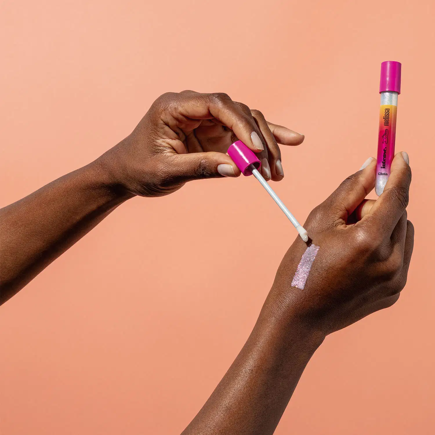 Uma mão feminina está segurando um tubo em tons de rosa, branco e laranja do gloss labial incolor Glossy Plastic Lips da marca Intense Melissa. A outra não está com a tampa e aplicador espalhando o produto na pele. O fundo da imagem é laranja.