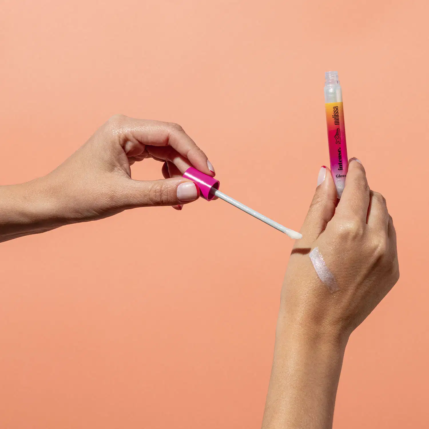 Uma mão feminina está segurando um tubo em tons de rosa, branco e laranja do gloss labial incolor Glossy Plastic Lips da marca Intense Melissa. A outra não está com a tampa e aplicador espalhando o produto na pele. O fundo da imagem é laranja.