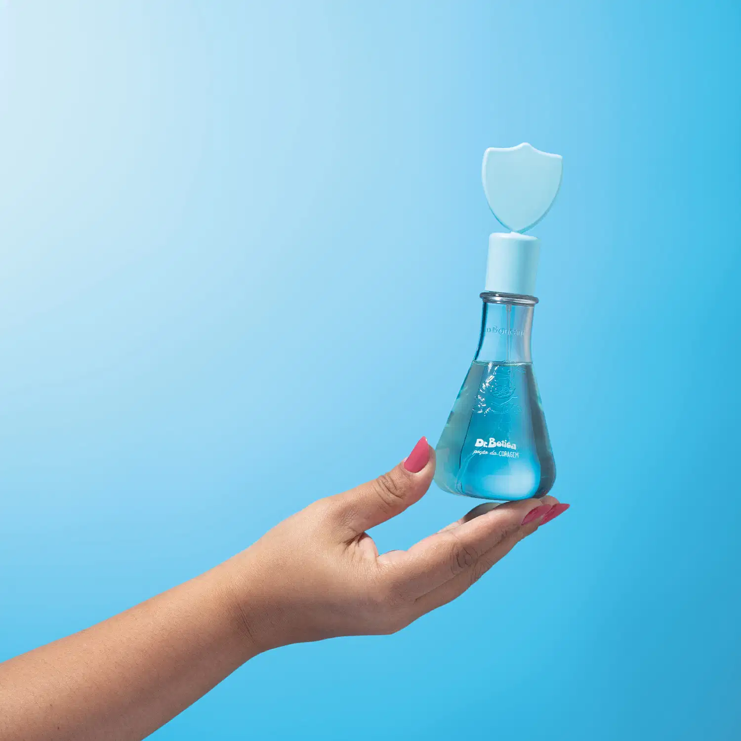 Mão feminina segurando um frasco transparente em formato de vidro laboratorial com líquido azul claro, tampa com escudo no topo da mesma tonalidade, contendo colônia infantil Poção da Coragem da marca Dr. Botica em fundo azul turquesa.