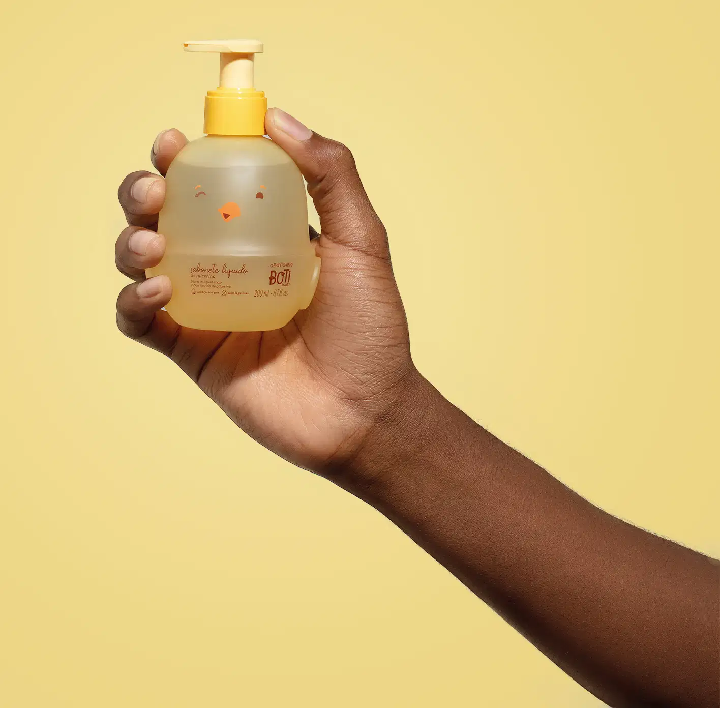 Mão segurando um frasco em forma de passarinho amarelo com detalhes laranjas contendo 200ml de sabonete líquido de glicerina da marca Boti Baby em um fundo alaranjado.