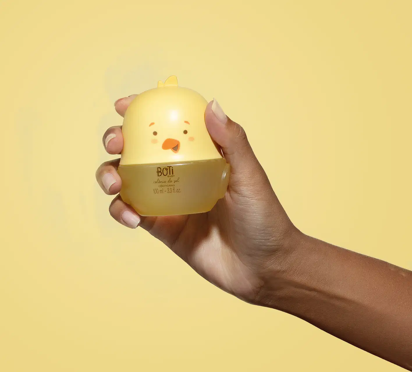 Mão segurando um frasco em formato de passarinho amarelo com parte superior na cor bege escuro e detalhes laranjas contendo 100ml de colônia do sol da marca Boti Baby em um fundo alaranjado.