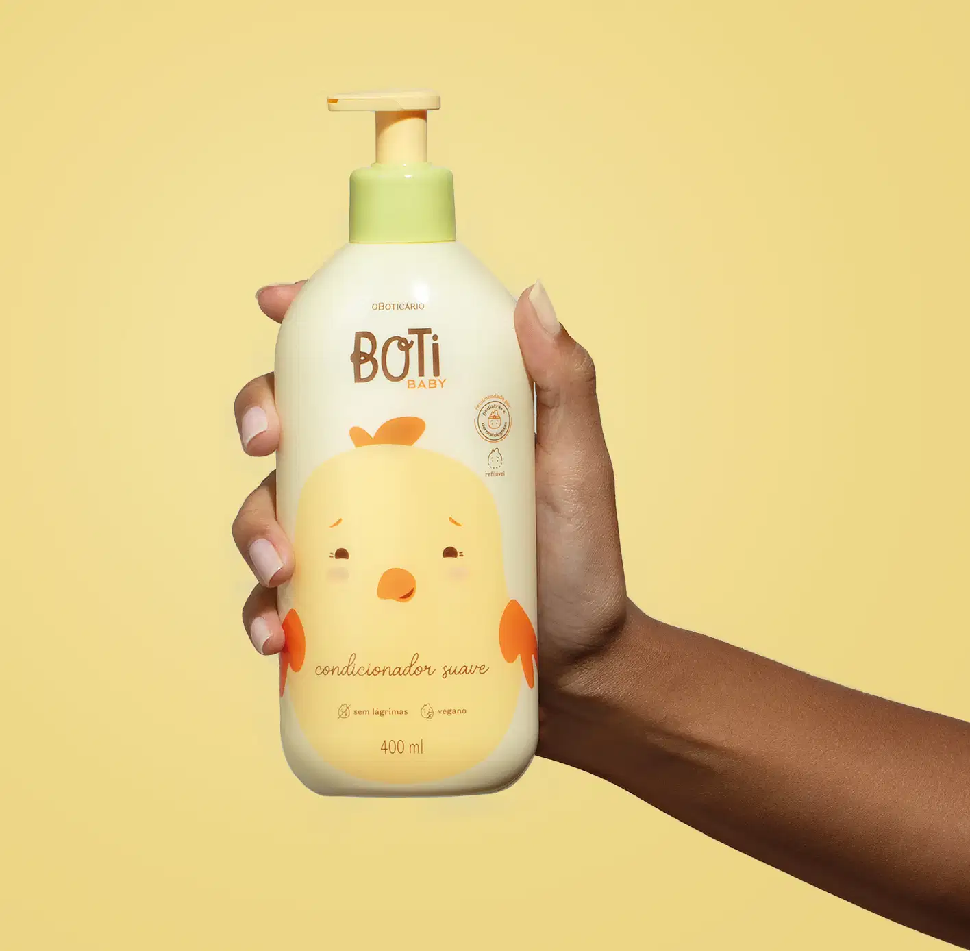 Mão segurando um frasco amarelo claro com ilustração de um passarinho amarelo com detalhes laranjas e bomba dosadora laranja de condicionador suave da marca Boti Baby em um fundo alaranjado.