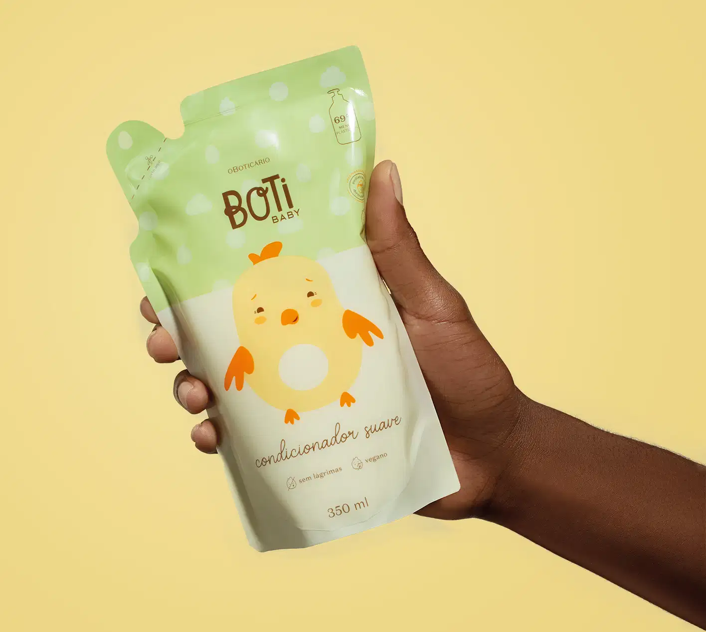 Mão segurando um refil de condicionador suave da marca Boti Baby em forma de sachê nas cores rosa e amarelo claro com ilustração de passarinho amarelo com detalhes laranjas em fundo alaranjado.