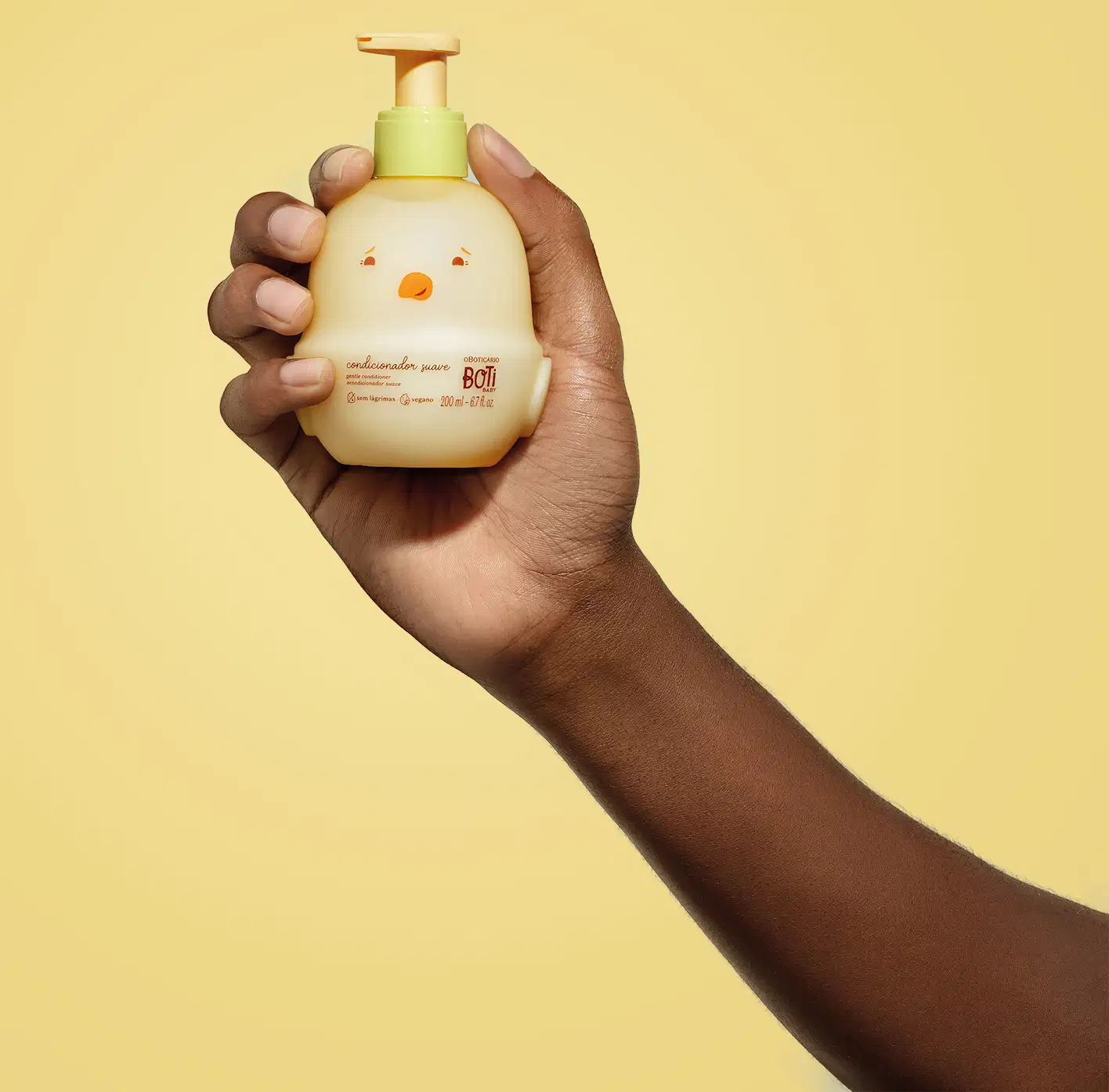 Mão segurando um frasco em forma de passarinho amarelo com detalhes laranjas contendo 200ml de condicionador suave da marca Boti Baby em um fundo alaranjado.