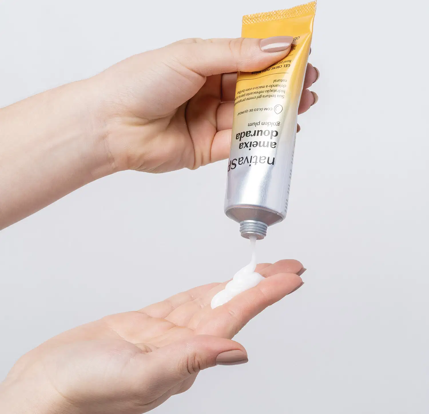 Uma mão está segurando uma bisnaga com degradê de prata e amarelo com letras pretas contendo gel creme para as mãos da marca Nativa SPA Ameixa Dourada e colocando um pouco do creme na outra mão. O fundo da imagem é azul claro.