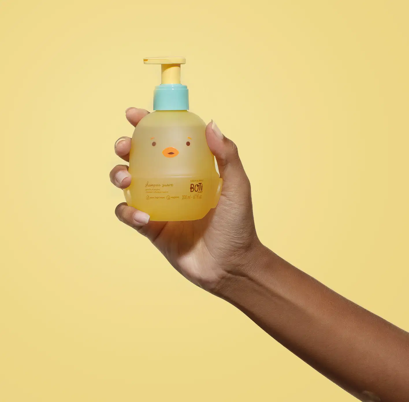Mão segurando um frascoem forma de passarinho amarelo com detalhes laranjas, tampa azul e bomba dosadora amarela de shampoo suave da marca Boti Baby em um fundo alaranjado.