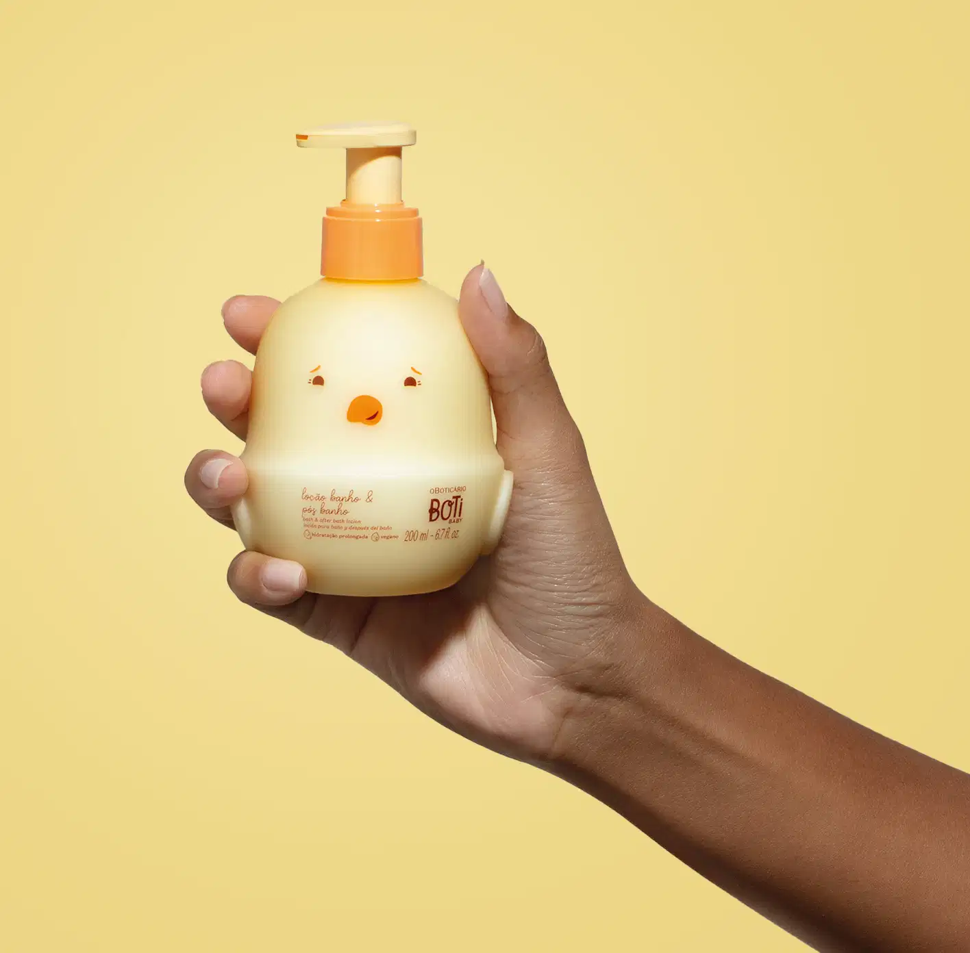 Mão segurando um frasco em forma de passarinho amarelo com detalhes laranjas contendo 200ml de loção hidratante banho e pós banho da marca Boti Baby em um fundo alaranjado.