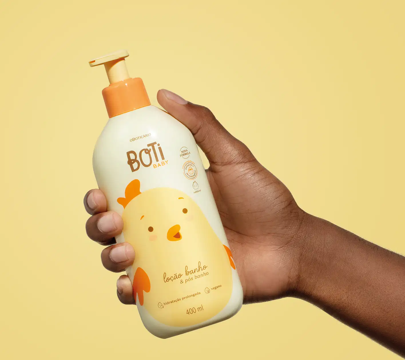 Mão segurando um frasco amarelo claro com ilustração de um passarinho amarelo com detalhes, tampa e bomba dosadora laranjas contendo 400ml de loção hidratante banho e pós banho da marca Boti Baby em um fundo alaranjado.