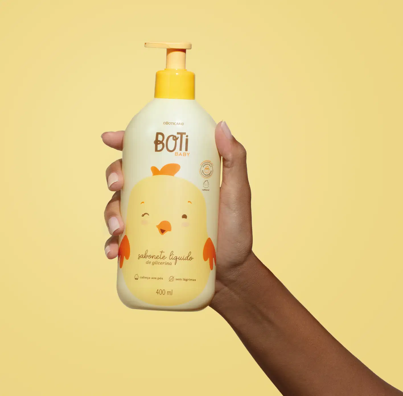 Mão segurando um frasco amarelo claro com ilustração de um passarinho amarelo com detalhes, tampa e bomba dosadora laranjas contendo sabonete líquido de glicerina da marca Boti Baby em um fundo alaranjado.